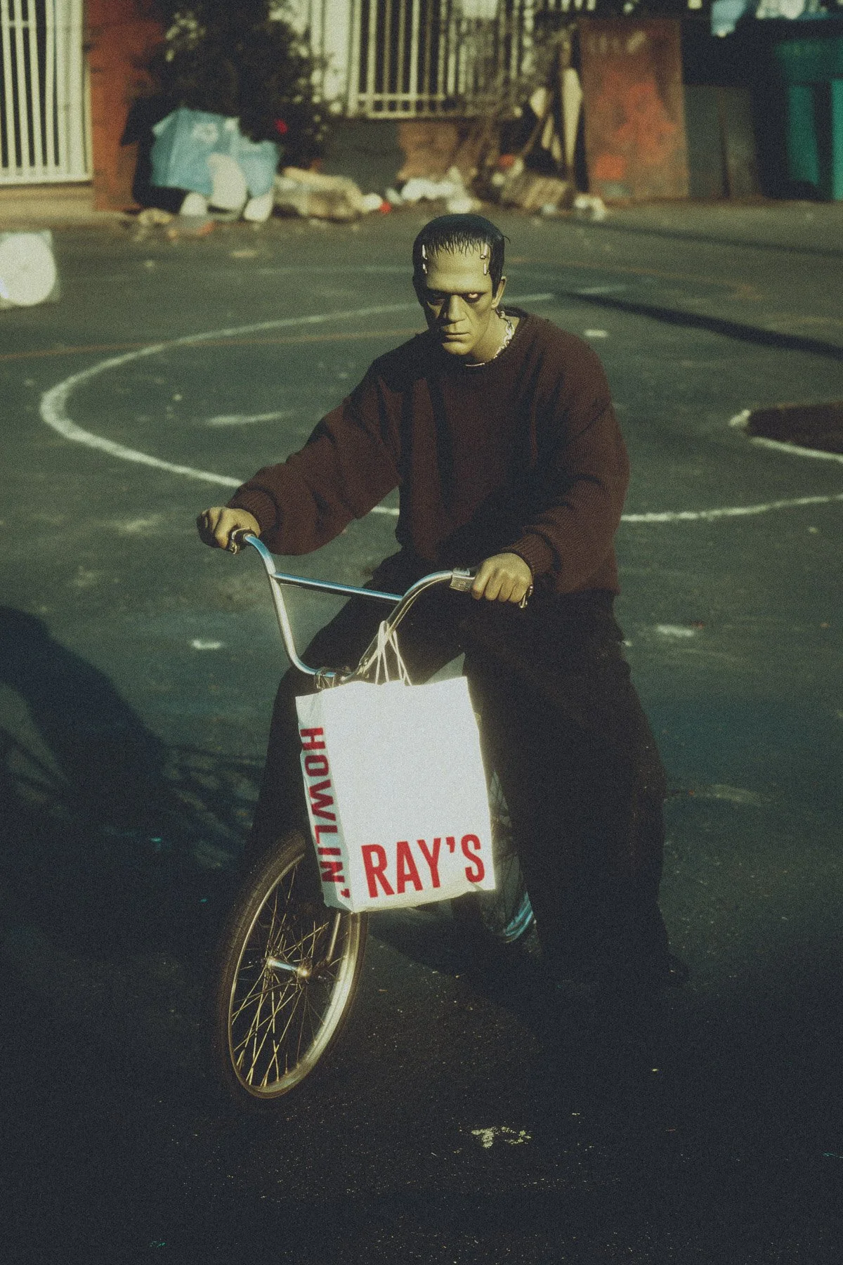 A person with dark hair and a serious expression riding a bicycle on a street at night. The bicycle has a white basket with red lettering reading 'RAY'S' and other words partially visible. The background includes a trash can, a gated staircase, and a