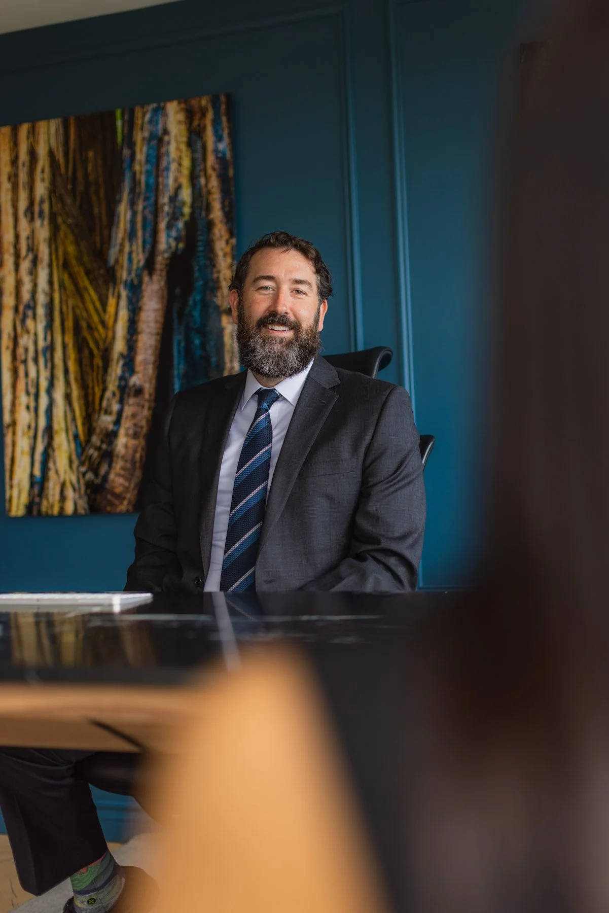 A man with a beard and mustache in a gray suit and tie sitting in an office with teal walls and colorful artwork behind him.