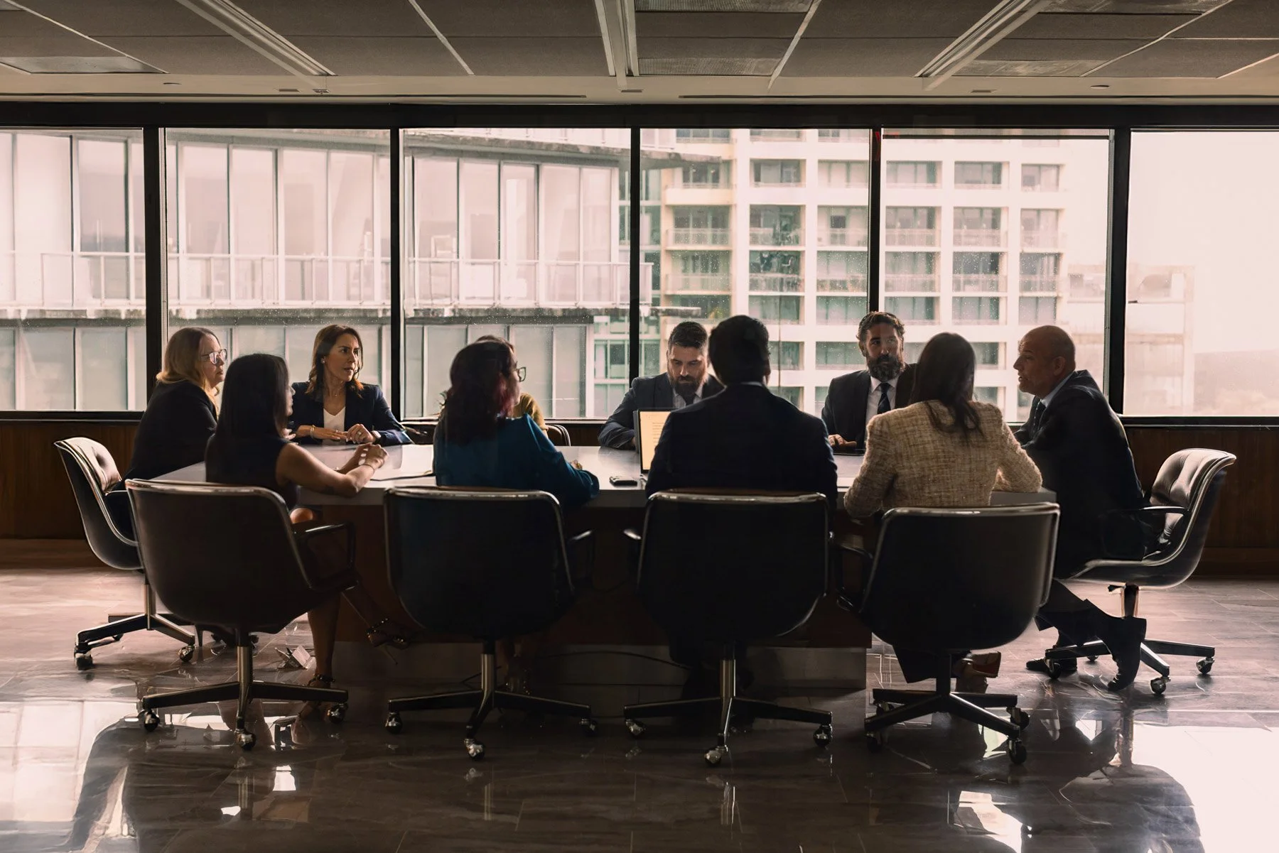 A business meeting taking place in a conference room with large windows, showing people seated around a circular table engaged in discussion.