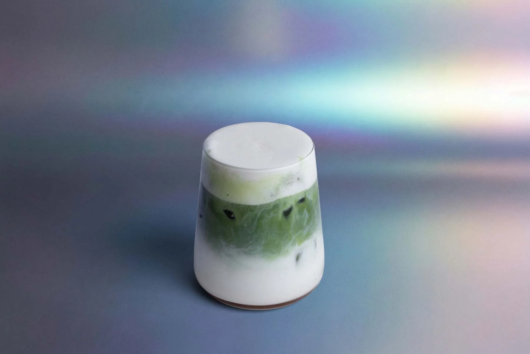 A glass of green matcha latte topped with foam, set against a background with a rainbow gradient
