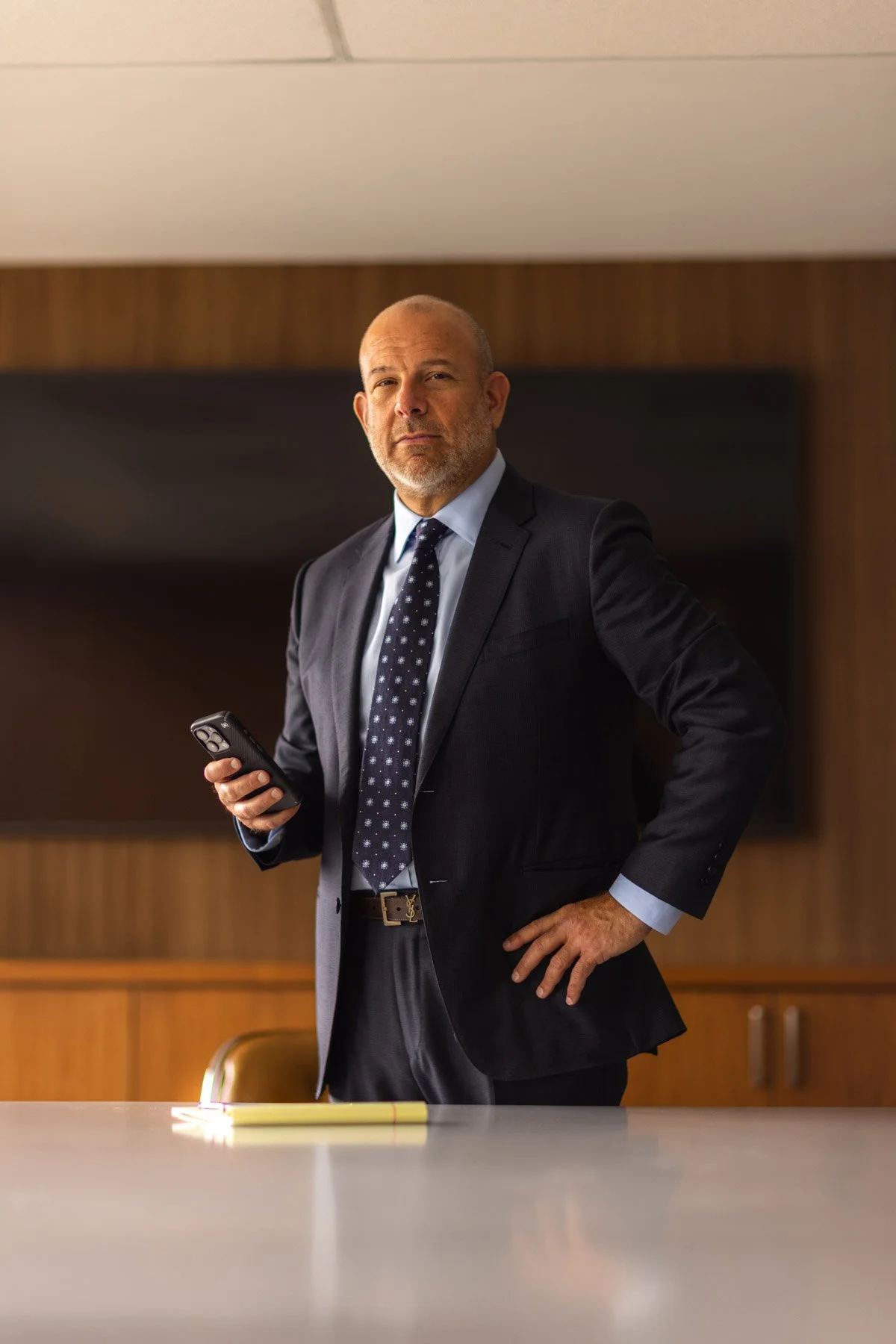 A middle-aged man in a dark suit, light blue shirt, and patterned tie standing in a conference room holding a smartphone in his right hand, with a serious expression on his face.
