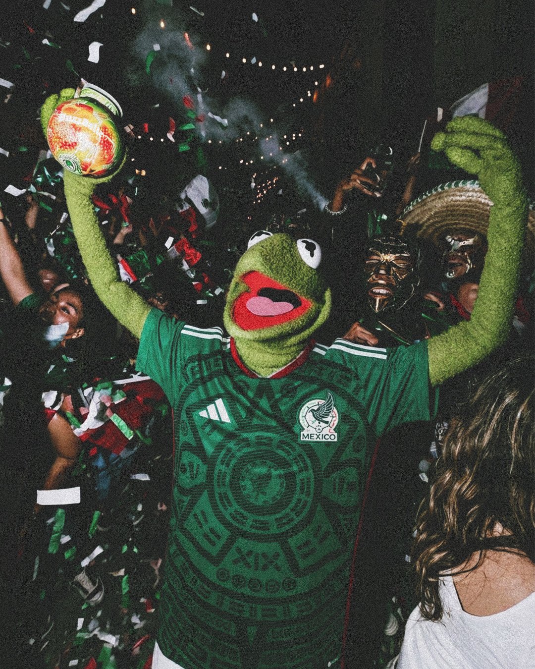 Kermit the frog in a green Mexico soccer jersey wearing a Pepe the Frog costume, holding a soccer ball, celebrating at a crowded event with confetti and party atmosphere at night.