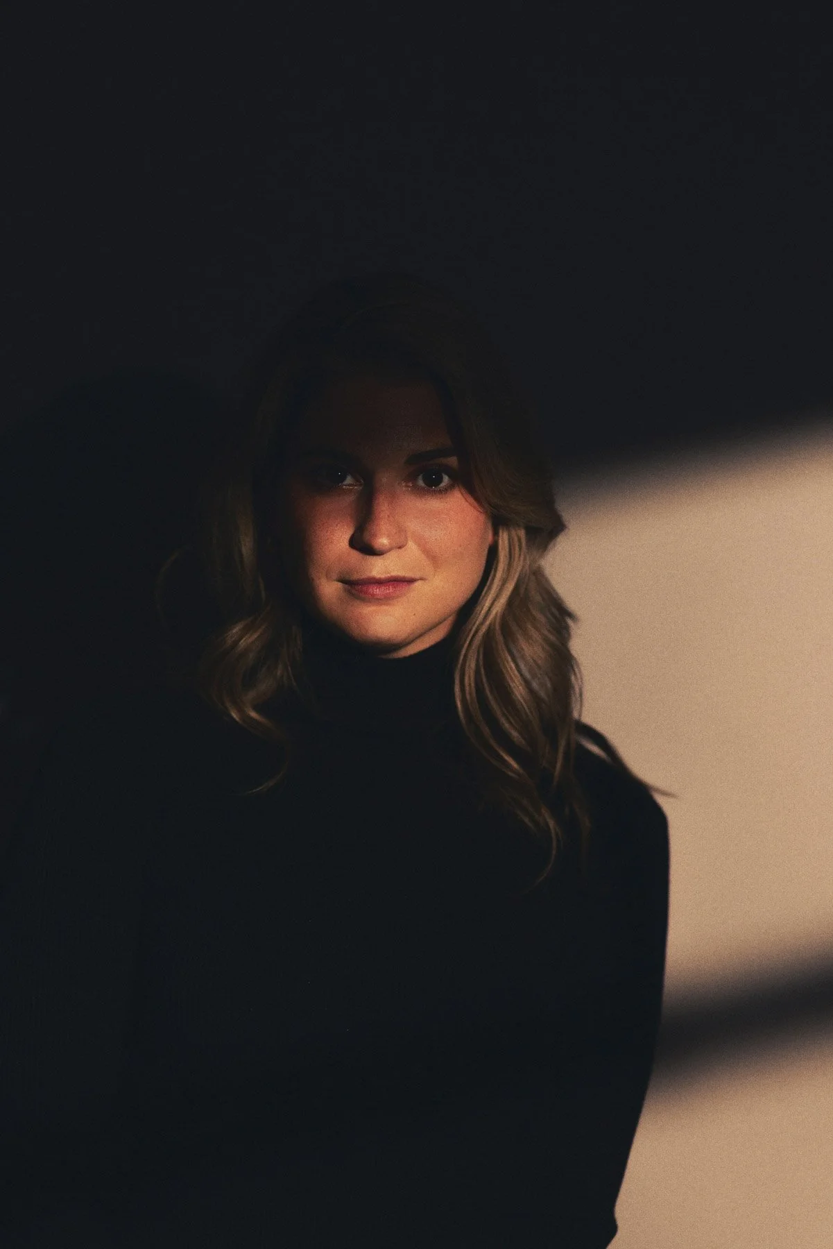 A woman with wavy hair is wearing a black turtleneck and looking directly at the camera with a subtle smile. The background is dark with a gradient light on the right side.