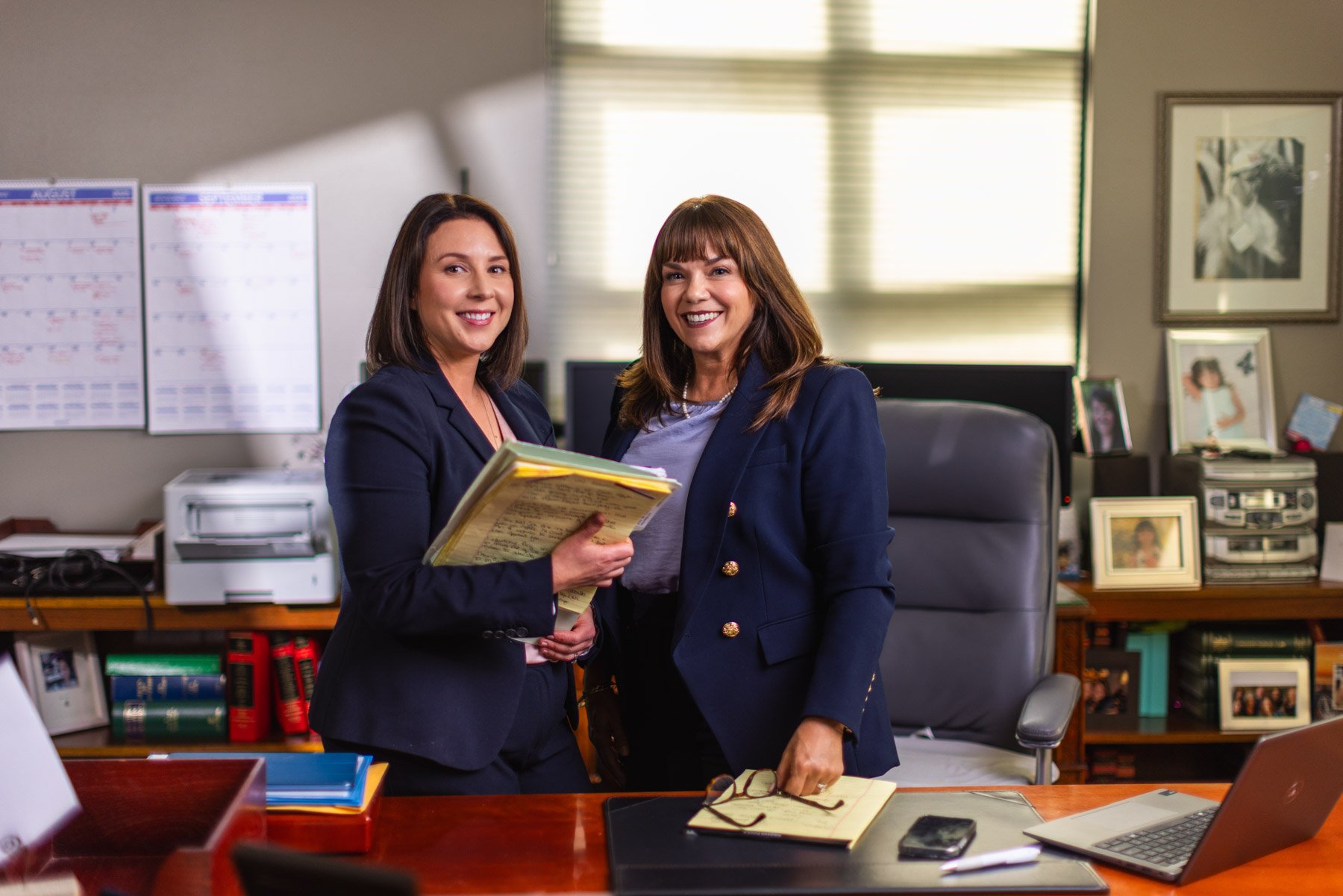 Two women in business attire standing in an office, smiling at the camera, one holding a notepad. The office has shelves with books, framed photos, and various office supplies, with sunlight coming through window blinds.