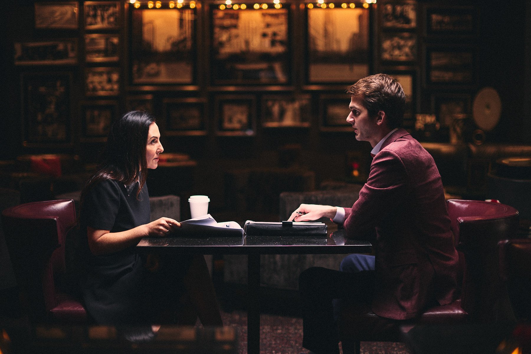 A man and a woman are sitting across from each other at a table in a dimly lit restaurant or cafe. They are engaged in a serious conversation, with the woman holding some papers and the man taking notes on a notepad. There are framed pictures on the 