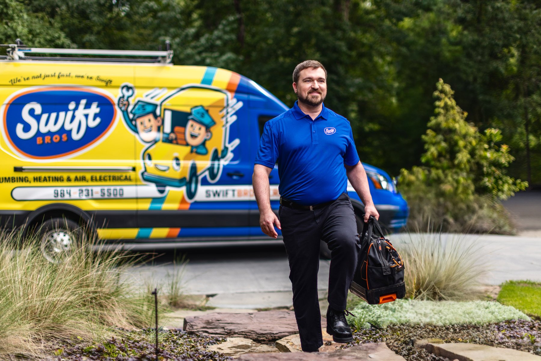 A man dressed in a blue company shirt walking on a stone pathway, carrying a black and orange tool bag, with a brightly colored Swift Bros service van and green trees in the background.