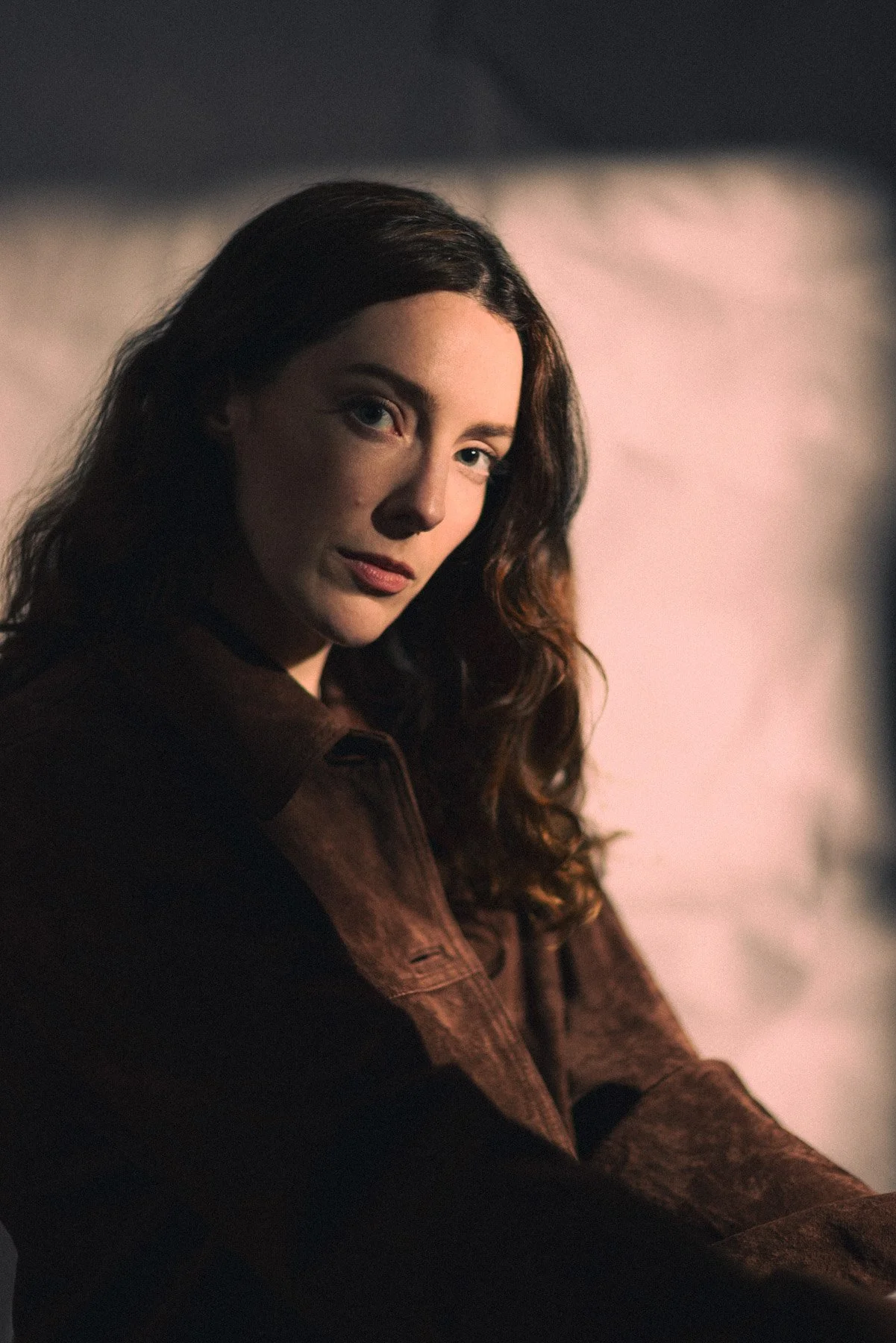 A woman with long curly brown hair, wearing a brown jacket, is sitting in a dimly lit room with a serious expression, looking toward the camera.