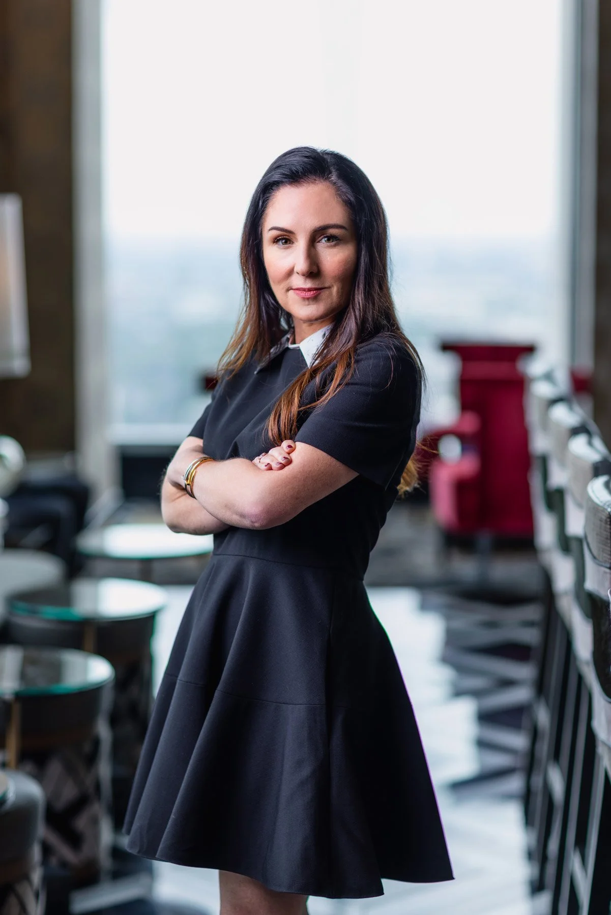 A woman with long dark hair, arms crossed, wearing a black dress, standing in a room with chairs and large windows in the background.