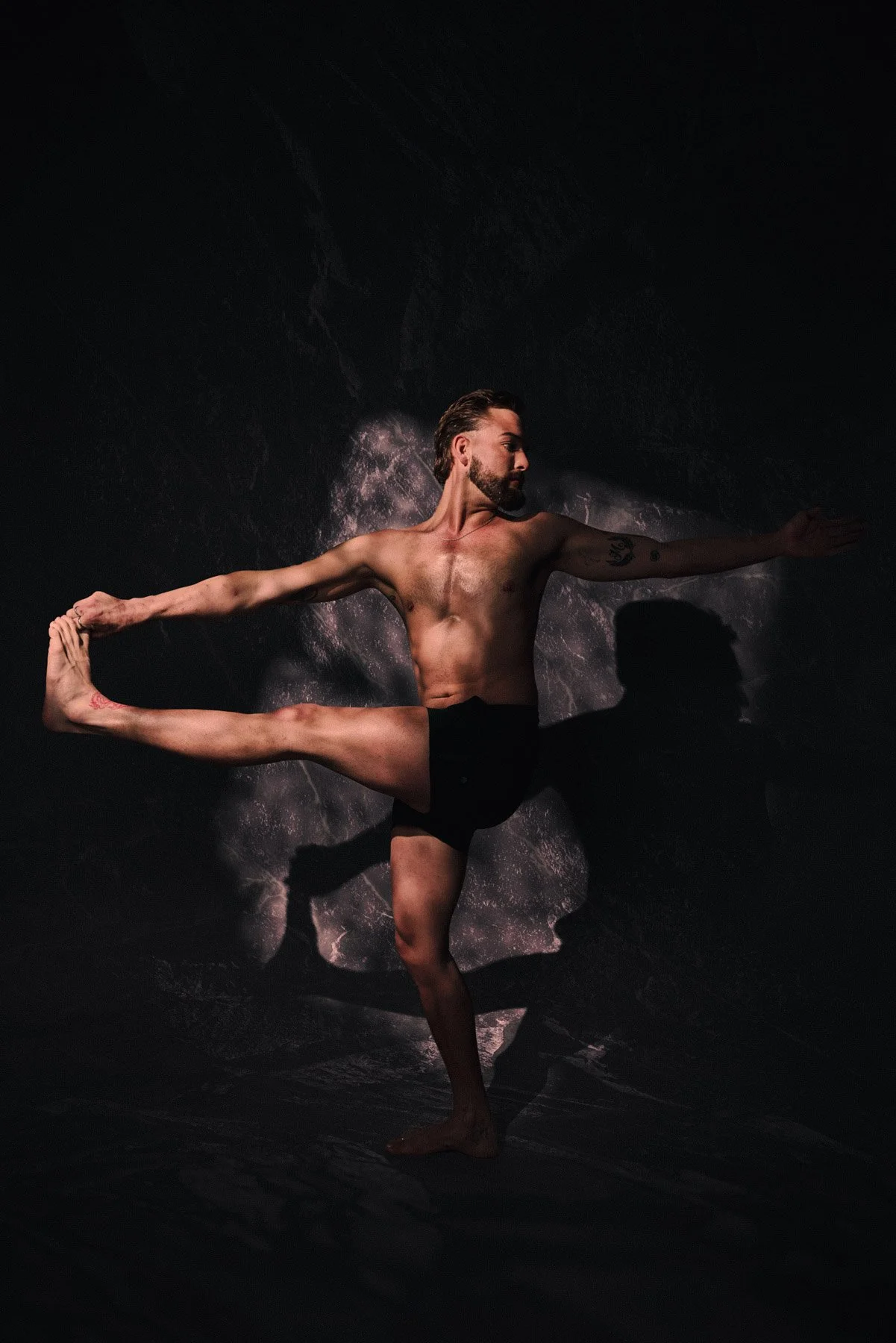 A shirtless man with tattoos performing a yoga pose against a dark background with subtle lighting.
