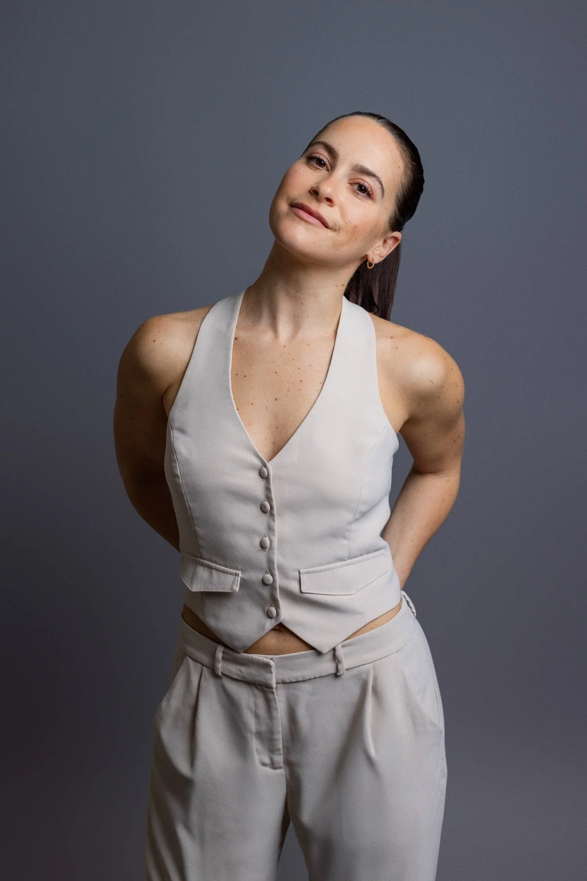 A woman with dark hair tied back, wearing a sleeveless beige vest with buttons and matching beige pants, standing against a dark gray background. She has a slight smile and looks straight at the camera.