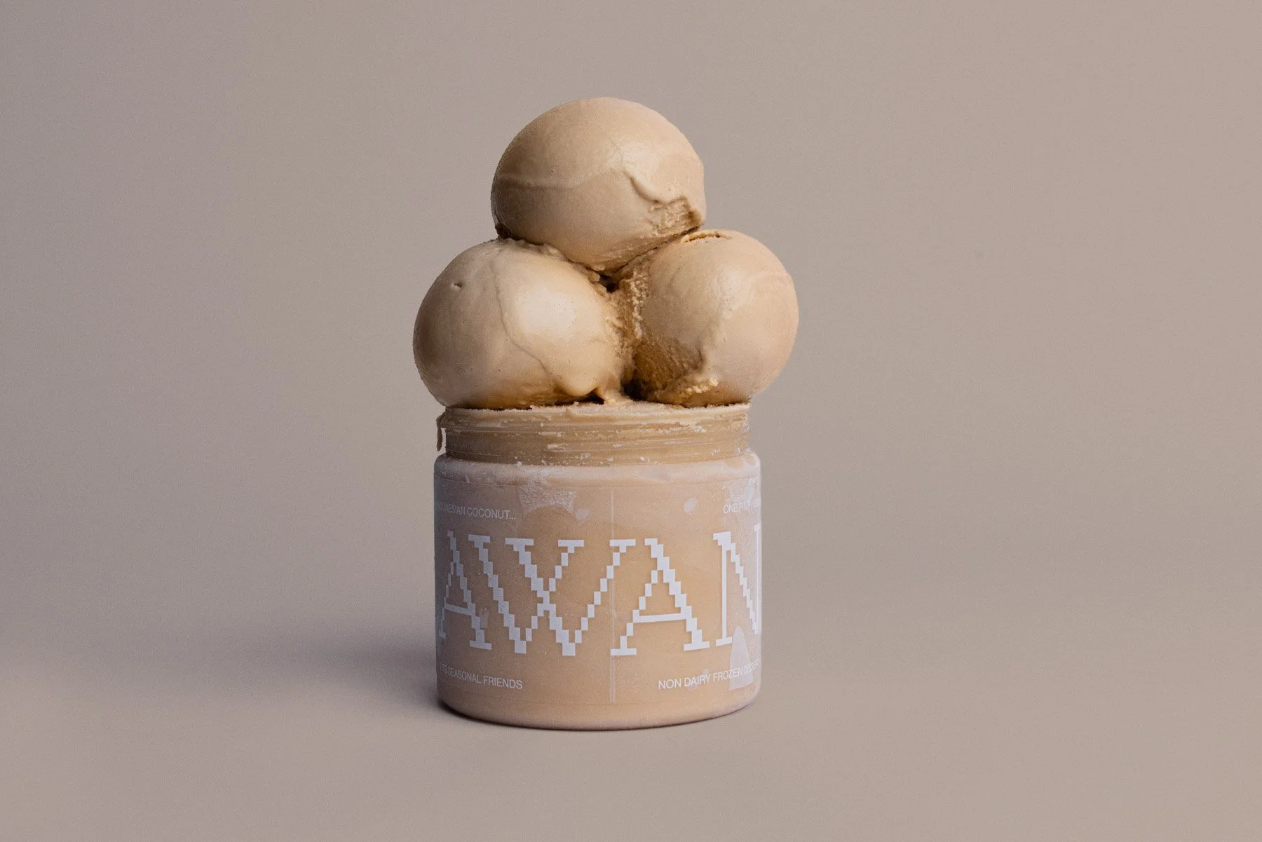 Three scoops of beige ice cream stacked on top of a container with a white and beige label, against a plain neutral background.
