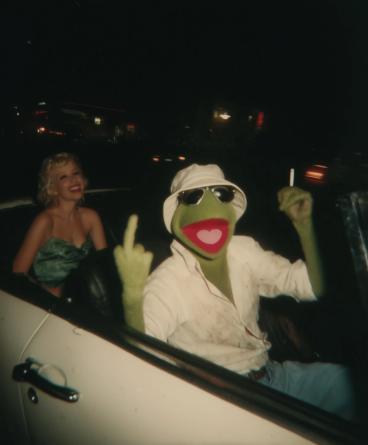 A woman and a frog puppet sitting in a car at night, with the woman smiling and the frog wearing sunglasses, a hat, and a white shirt, with its tongue showing and making a gesture.