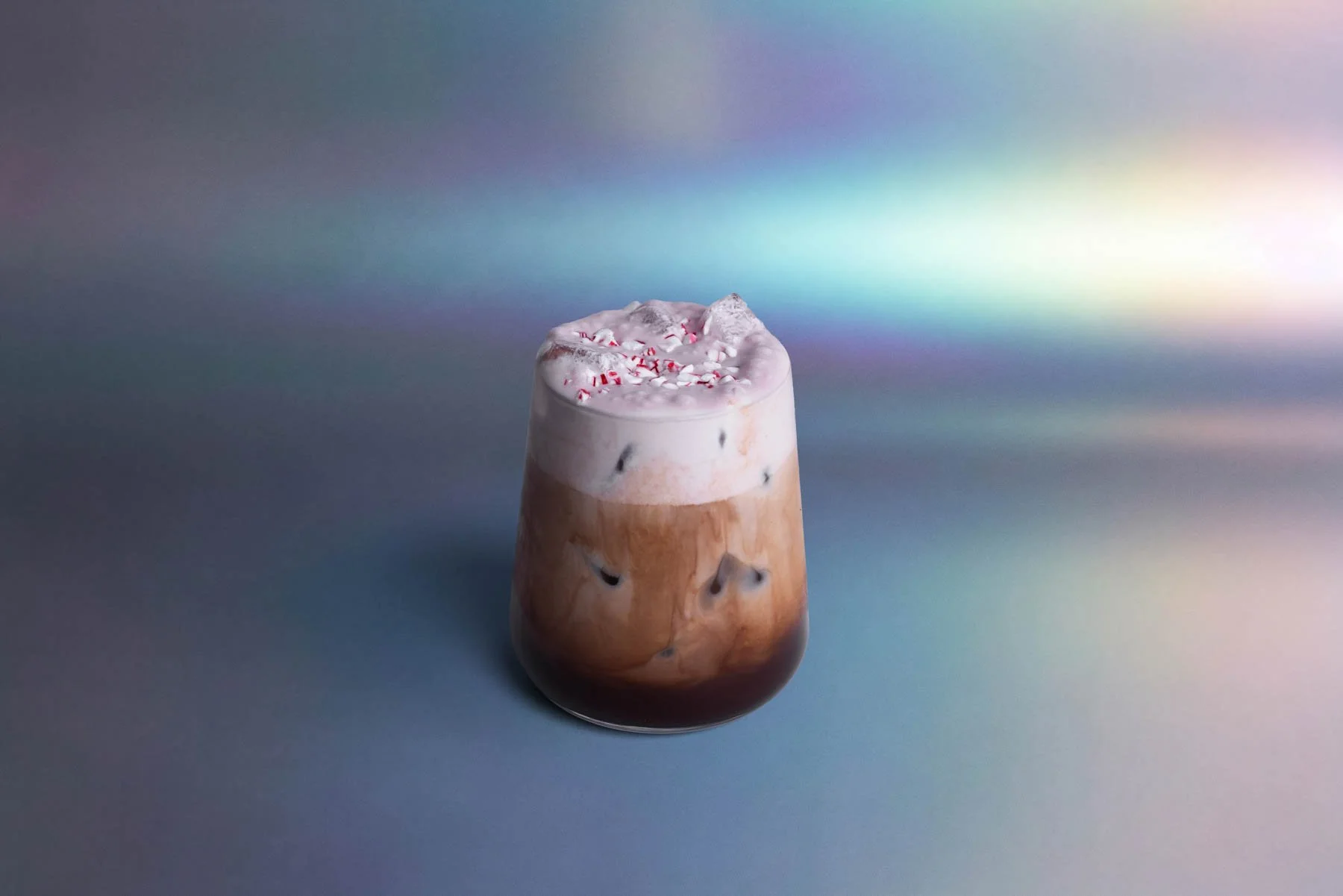 A glass filled with a layered iced coffee drink topped with whipped cream and red and white peppermint candy pieces, set against a holographic background.
