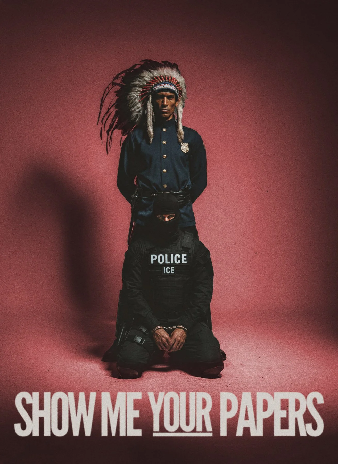 An art installation featuring a man wearing a Native American headdress and police gear, with the caption 'SHOW ME YOUR PAPERS' at the bottom.