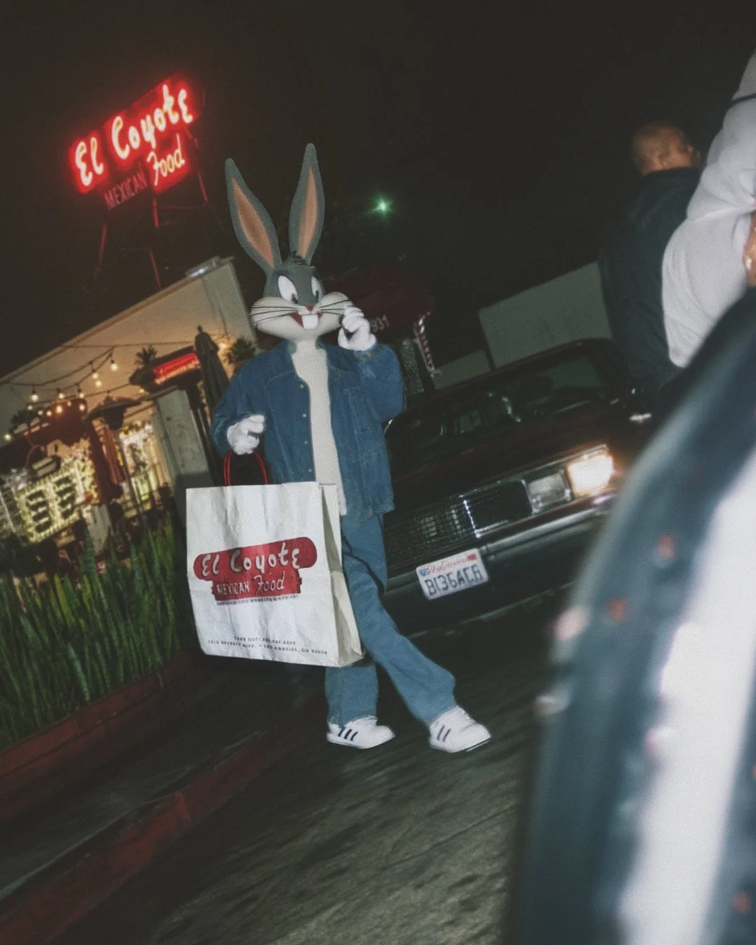 Bugs Bunny holding a shopping bag from El Coyote Mexican Food restaurant, standing outside at night near parked cars and a neon sign.