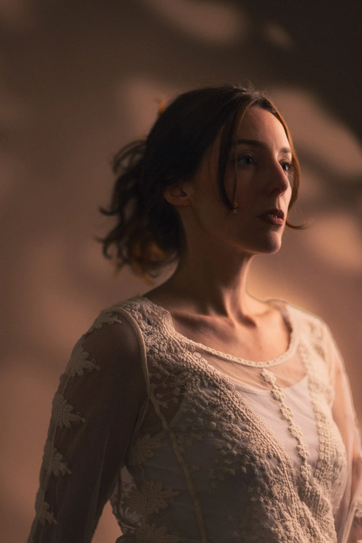 A woman with shoulder-length dark hair and a light-colored dress with lace details, looking to her right in a softly lit, neutral background.