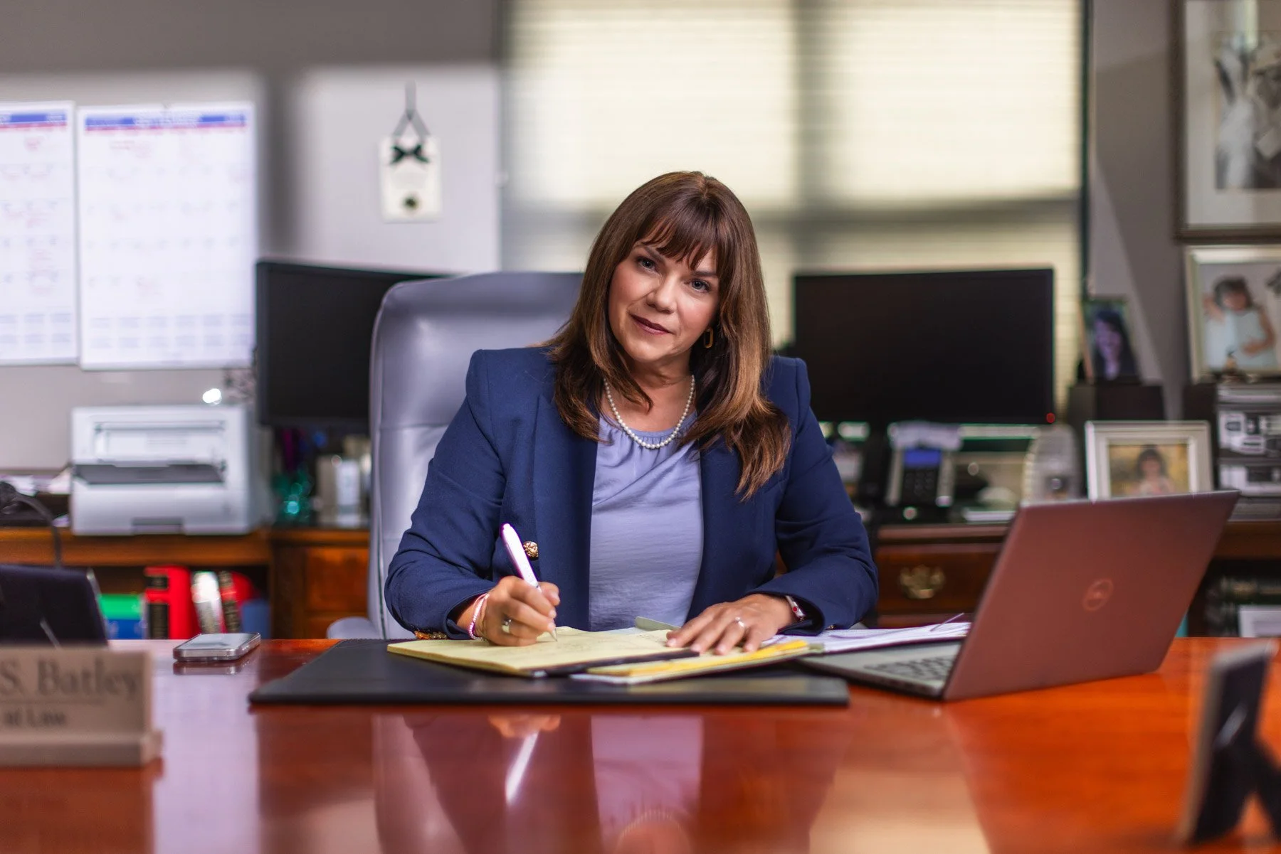 Woman in navy blazer and jewelry sitting at a large wooden desk in an office, writing on yellow notepad with a pen, with a silver laptop and office supplies on the desk.