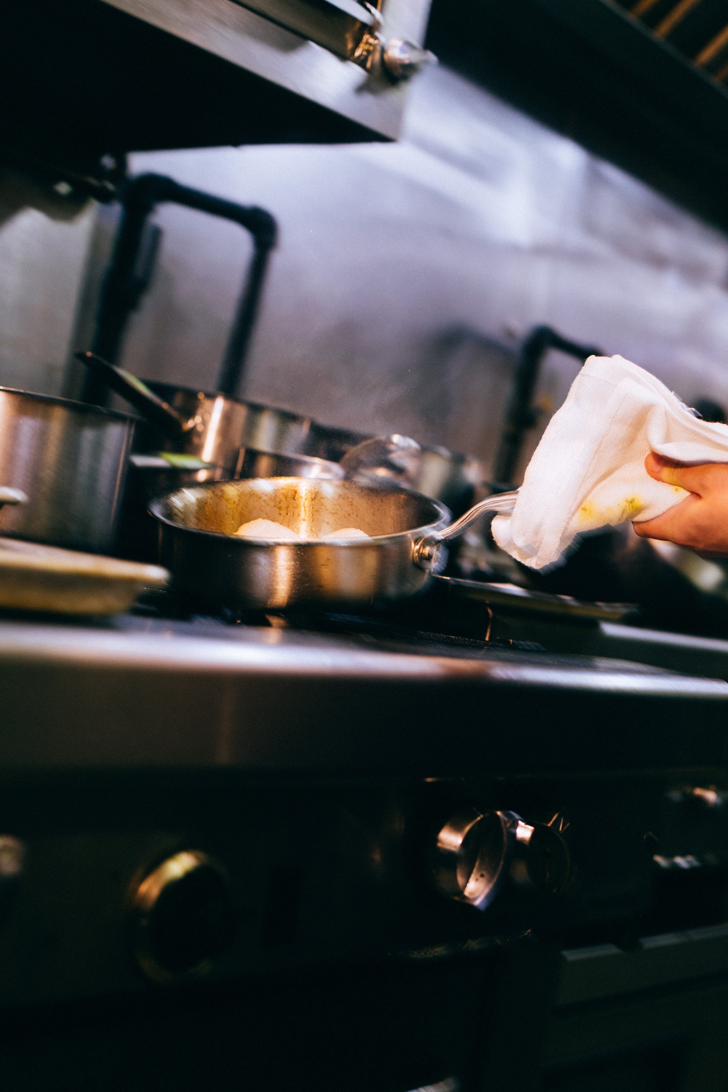 Person holding a frying pan with a cloth in a professional kitchen on a stove.