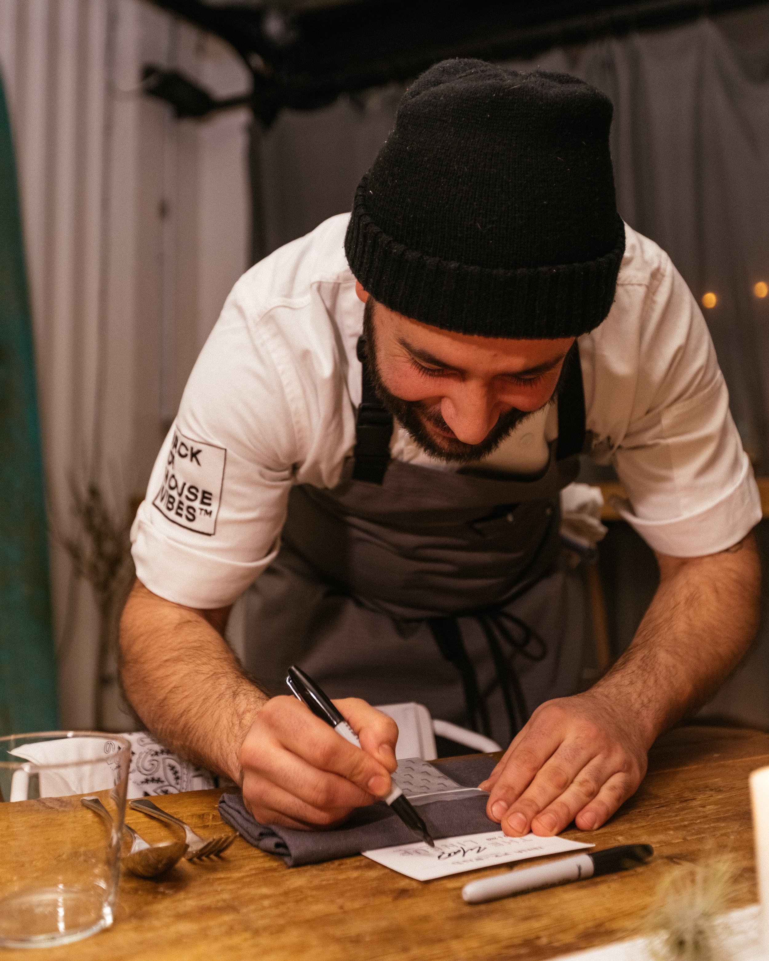 Chef writing on a note card at a wooden table, wearing a black beanie, apron, and white shirt.