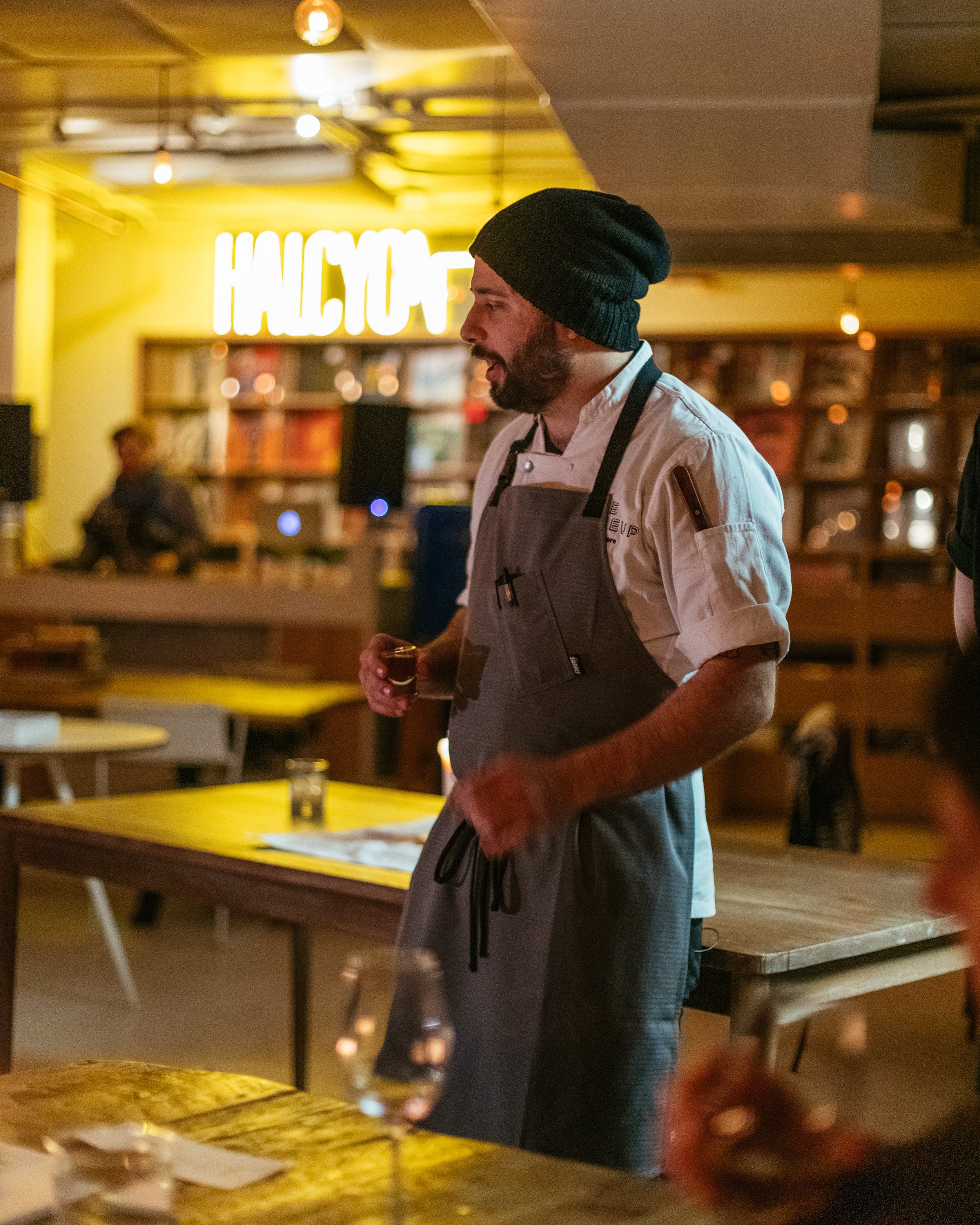 Chef wearing a beanie and apron speaking in a restaurant setting with yellow "HALCYON" sign in the background.