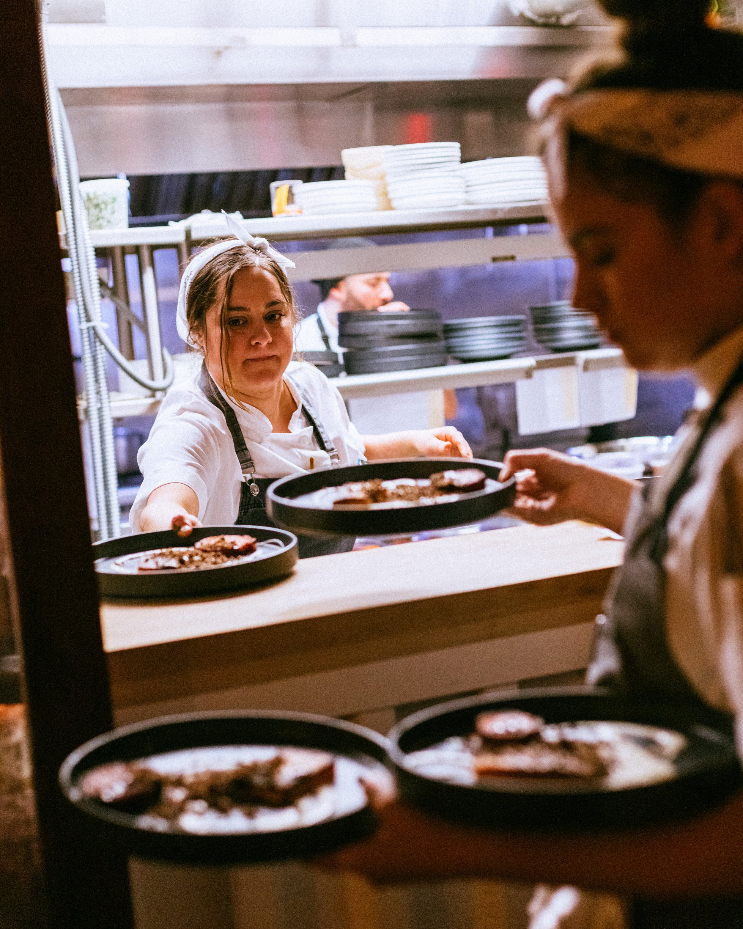 Two chefs in a restaurant kitchen, one handing plates to the other, with food preparation and dishes visible in the background.