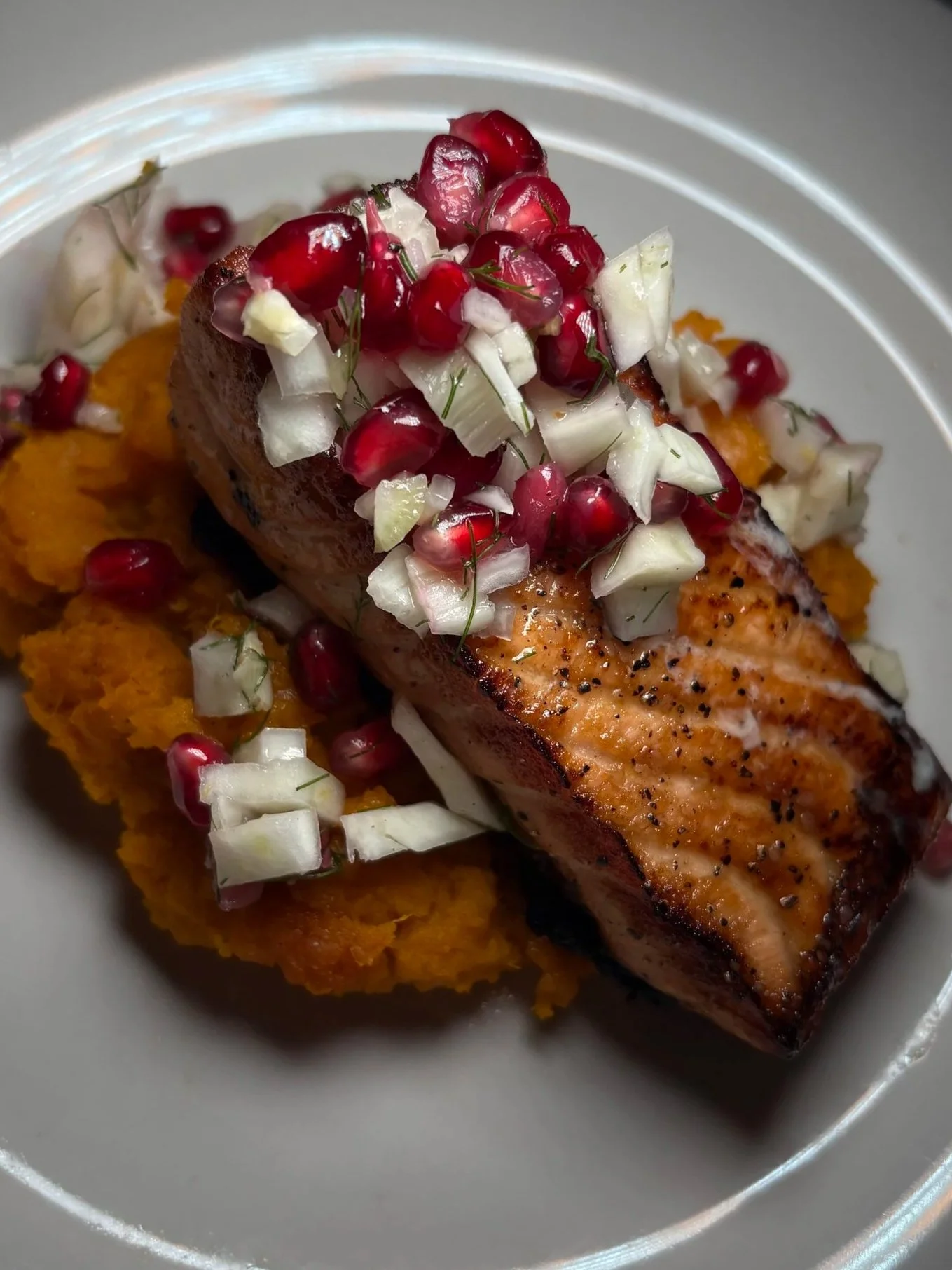 Cooked salmon fillet topped with chopped onions, pomegranate seeds, and herbs, served over mashed sweet potatoes.