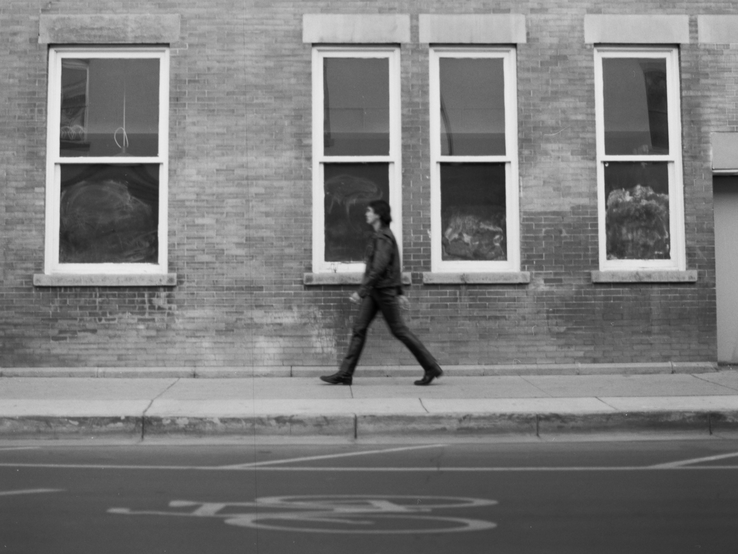 A person walking past a brick building with four windows, taken in black and white.