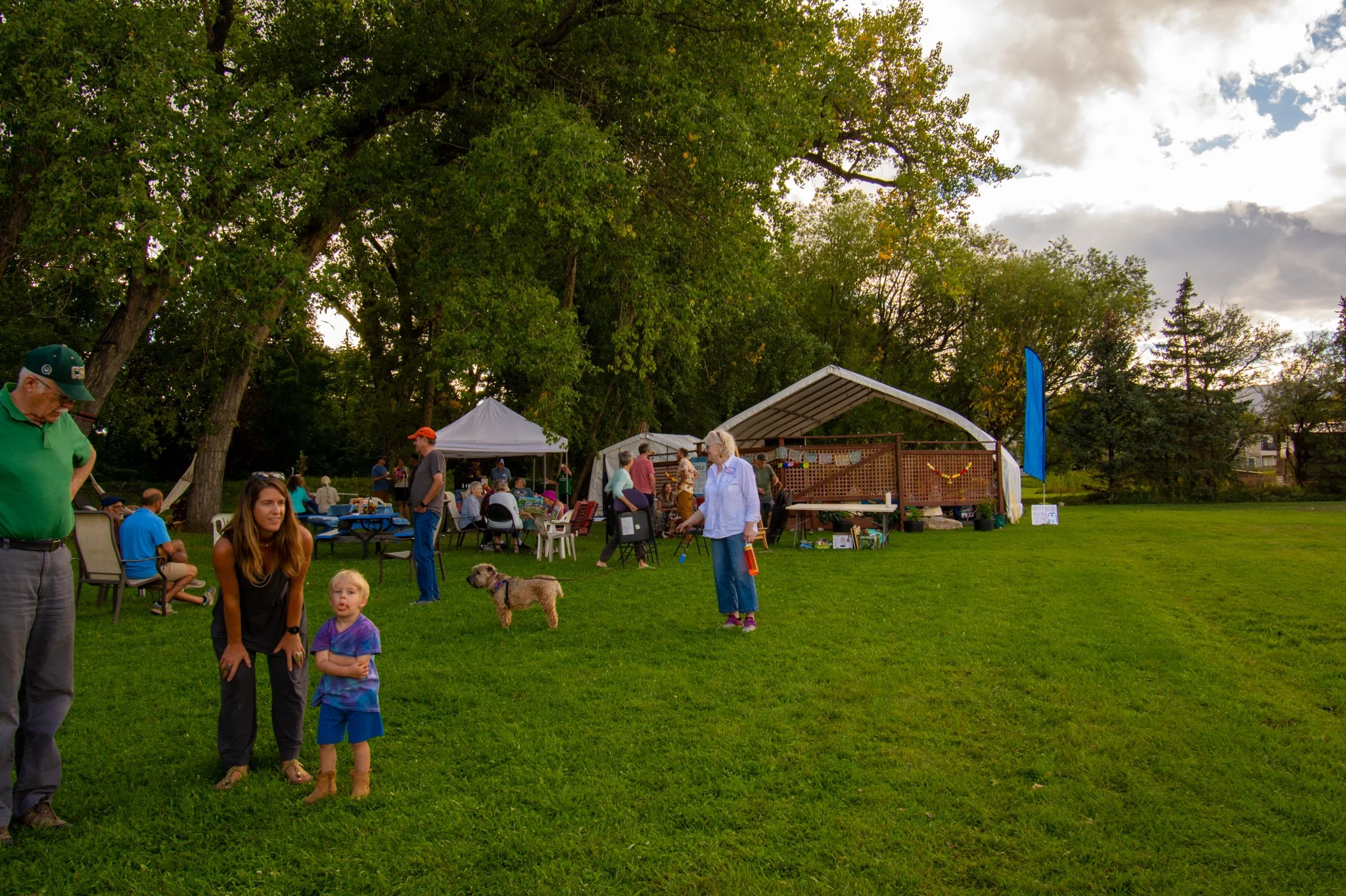 People gathered at an outdoor event in a grassy park with large trees, tents, and picnic tables under a partly cloudy sky.