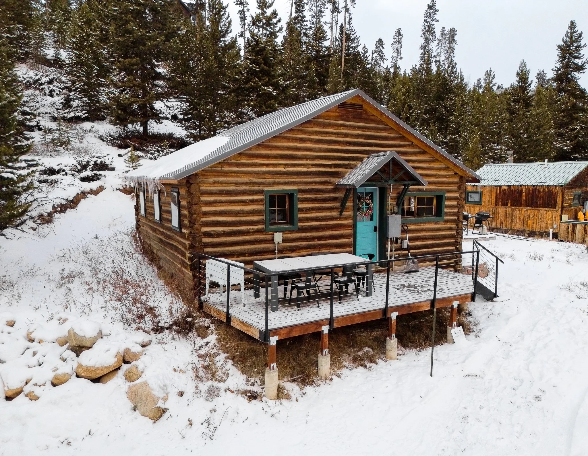 A small log cabin with a deck in a snowy, forested area, with pine trees in the background.