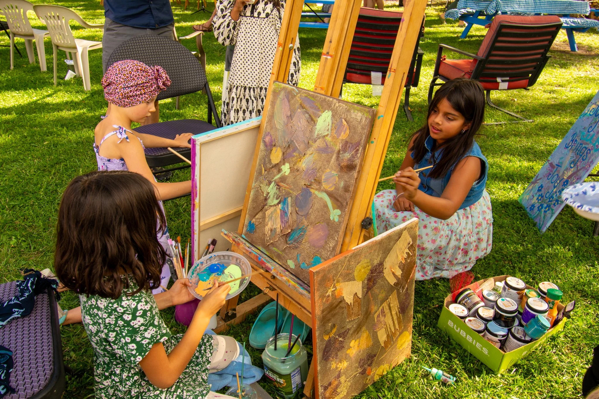 Three young girls painting on canvases outdoors on a grassy area with chairs and other paintings nearby.