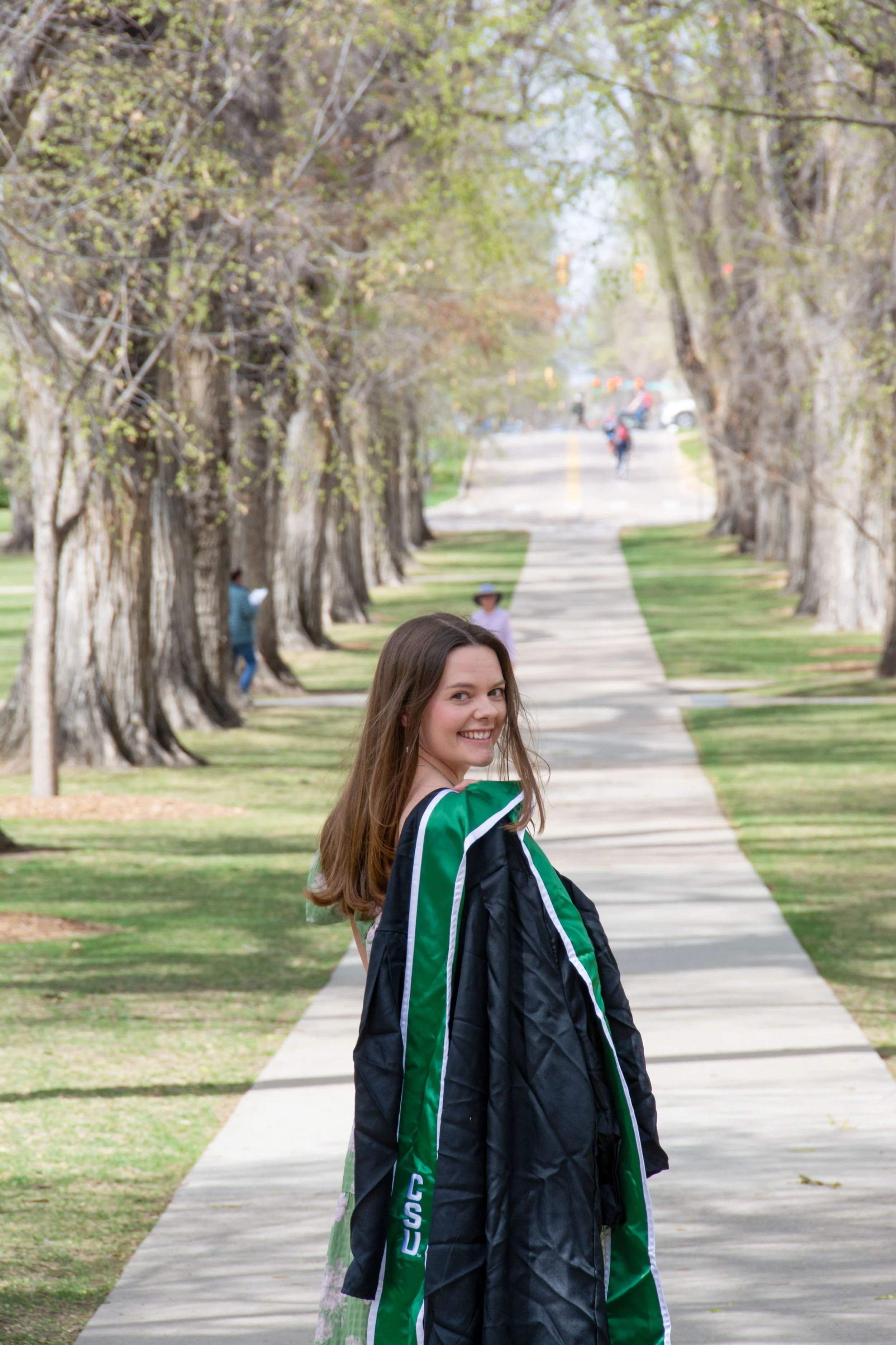 A smiling young woman with brown hair, wearing a graduation gown, standing on a tree-lined sidewalk in springtime.