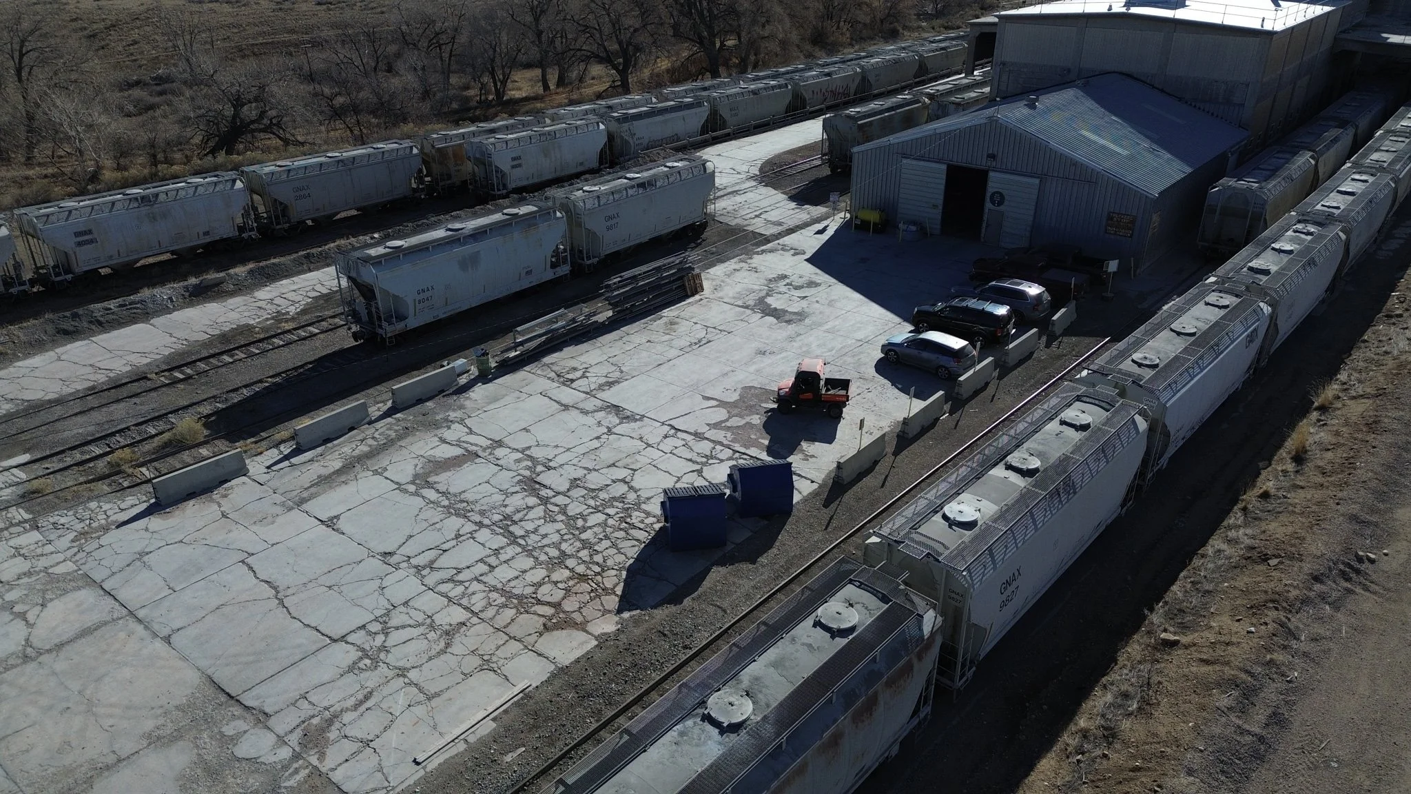 An aerial view of a train yard with multiple train cars parked along the tracks. There is a large gray building with a metal roof and some parked vehicles nearby, including a small red utility vehicle and several cars. The ground is cracked and weathered concrete, with some blue dumpsters and concrete barriers. Bare trees are visible in the background.