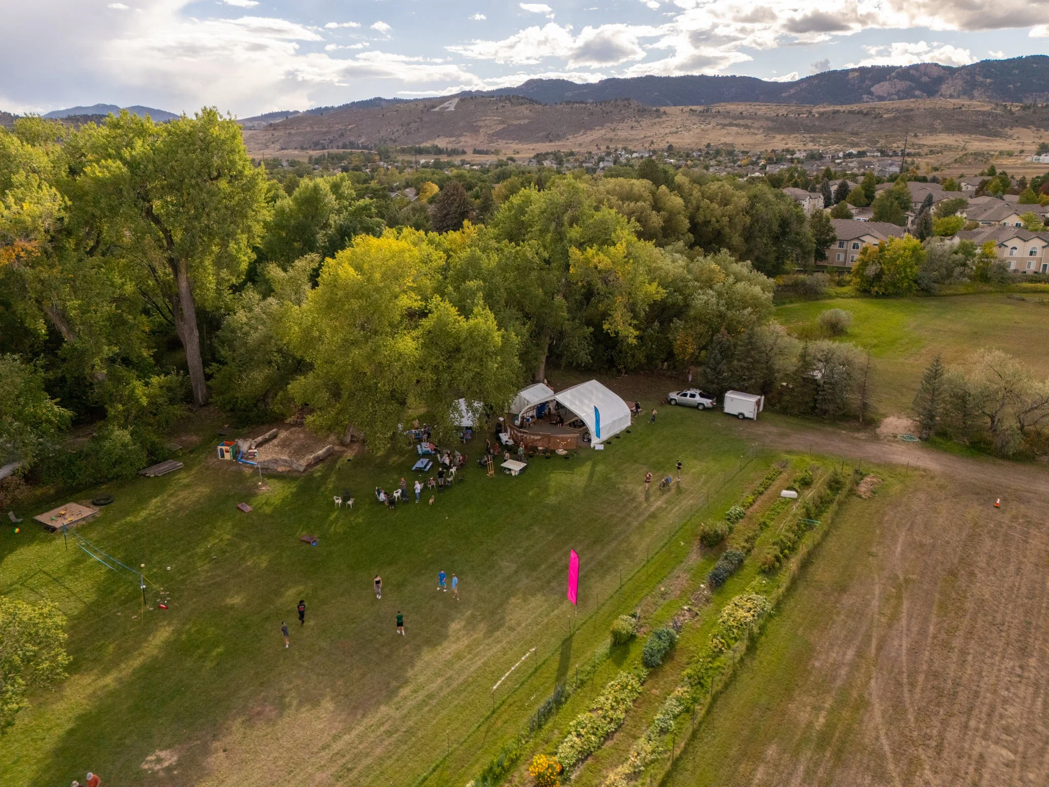Aerial view of a backyard gathering with tents, people, and outdoor activities, surrounded by trees and open fields, with mountain range in the background.