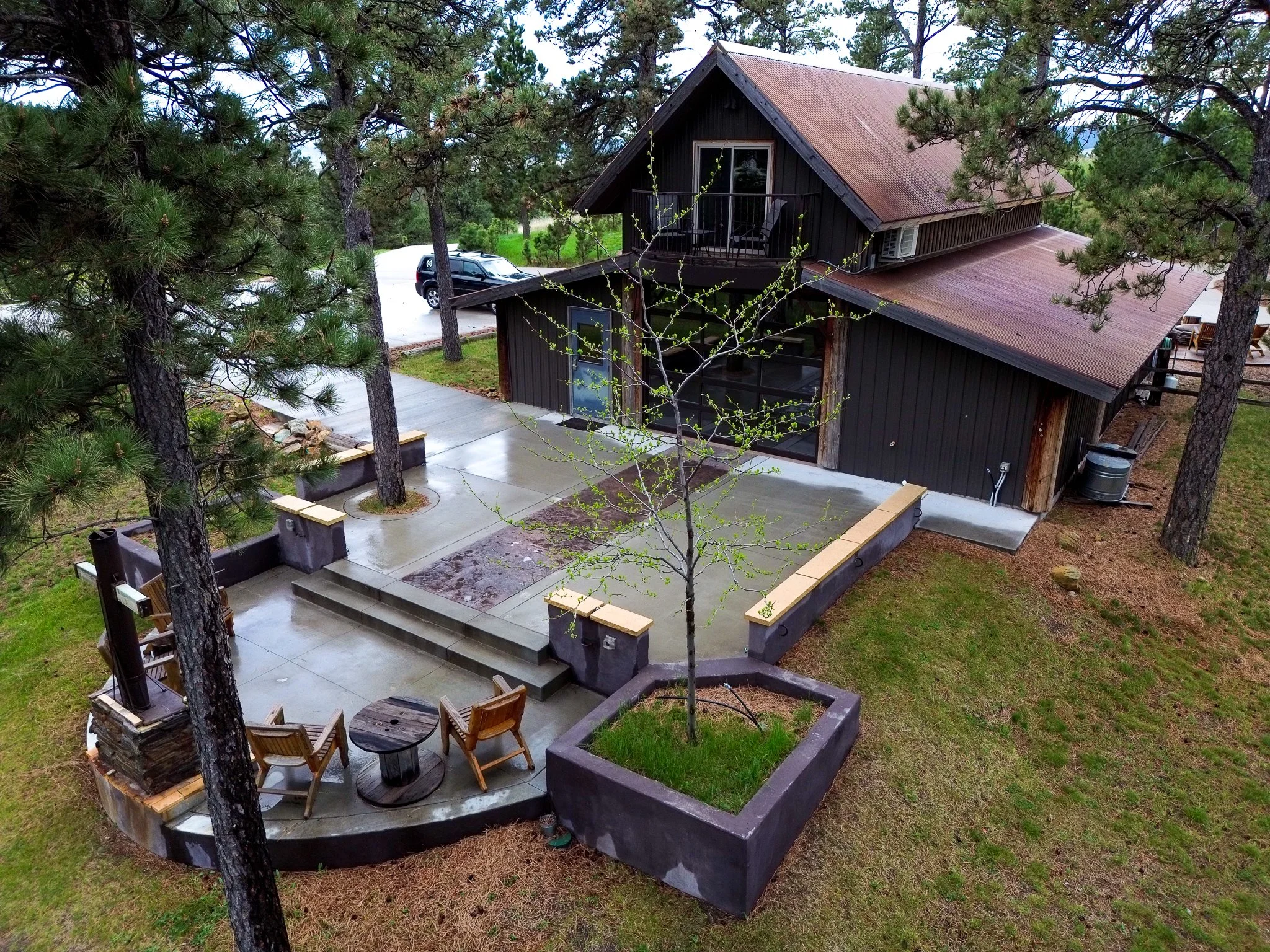 Aerial view of a black wooden house with a rust-colored metal roof, surrounded by trees and a large outdoor concrete patio with seating and a firepit.