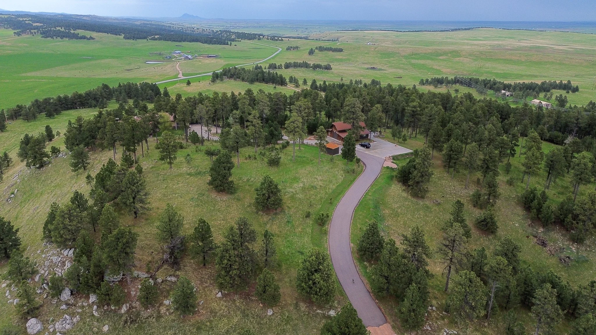 Aerial view of a rural property with a winding driveway leading to a house surrounded by trees and open fields, with more farmland and hills in the background.
