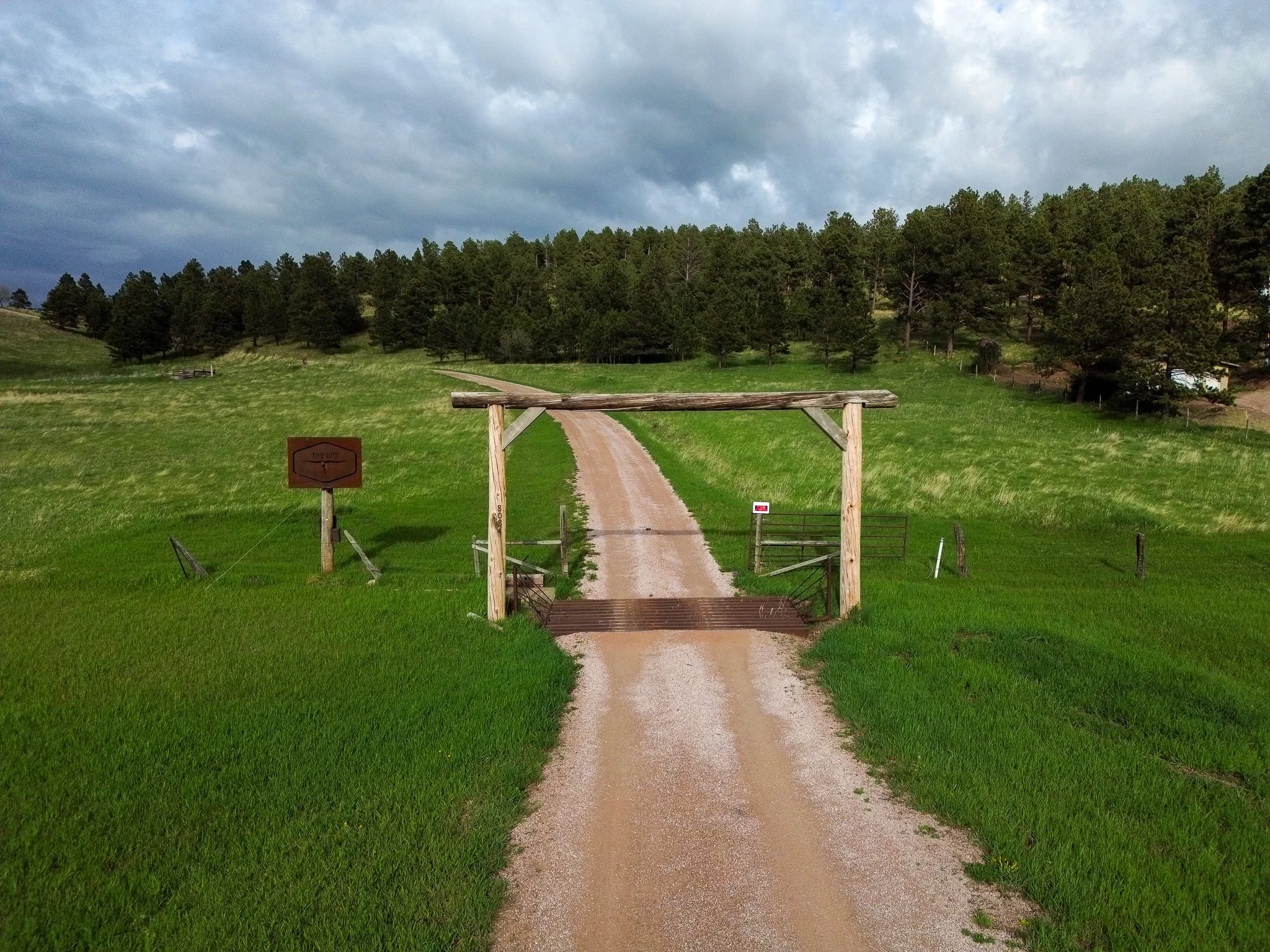 A dirt road leading through a wooden gate in a green field with trees in the background and cloudy sky overhead.
