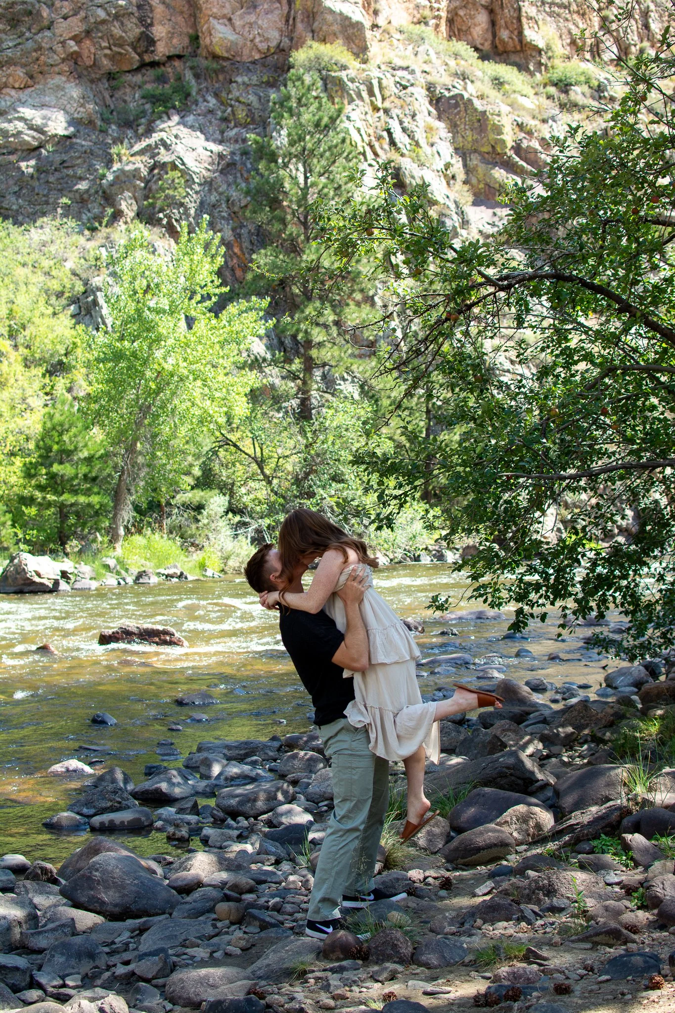 A man holding a woman in a dress by a river in a forested area with rocks and trees.