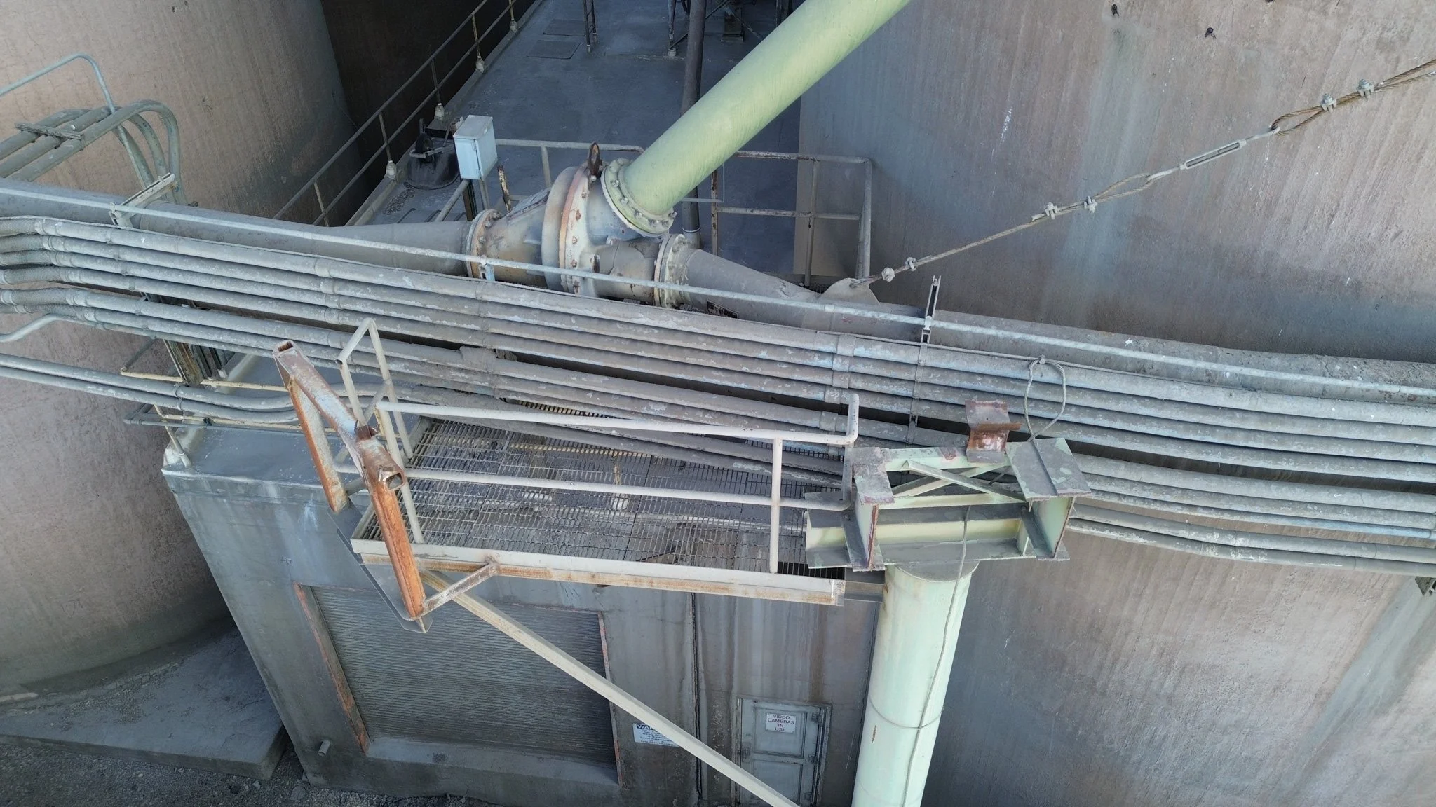 Industrial pipes and metal structures on a concrete wall, with a large green pipe connected to a smaller metal pipe with a flange, and some electrical wiring attached.