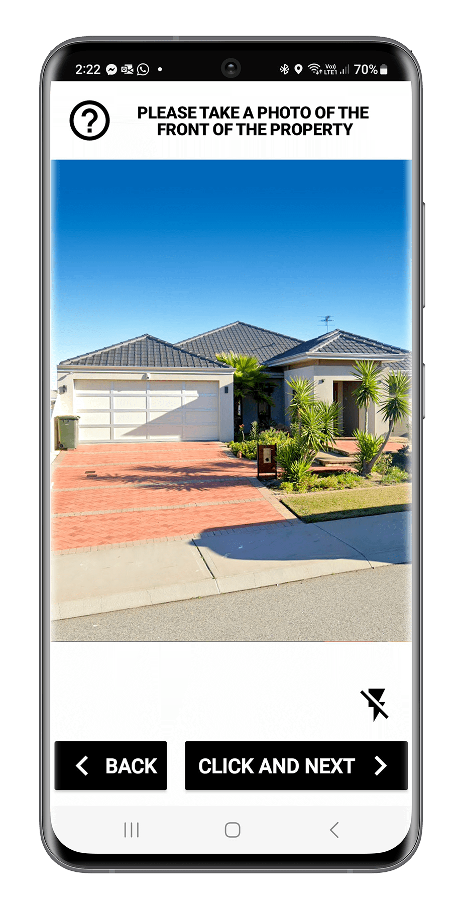A smartphone displaying an app screen asking to take a photo of the front of a house. The house has a gray tiled roof, white garage door, and a front yard with green plants and small trees. The sky is clear and blue.