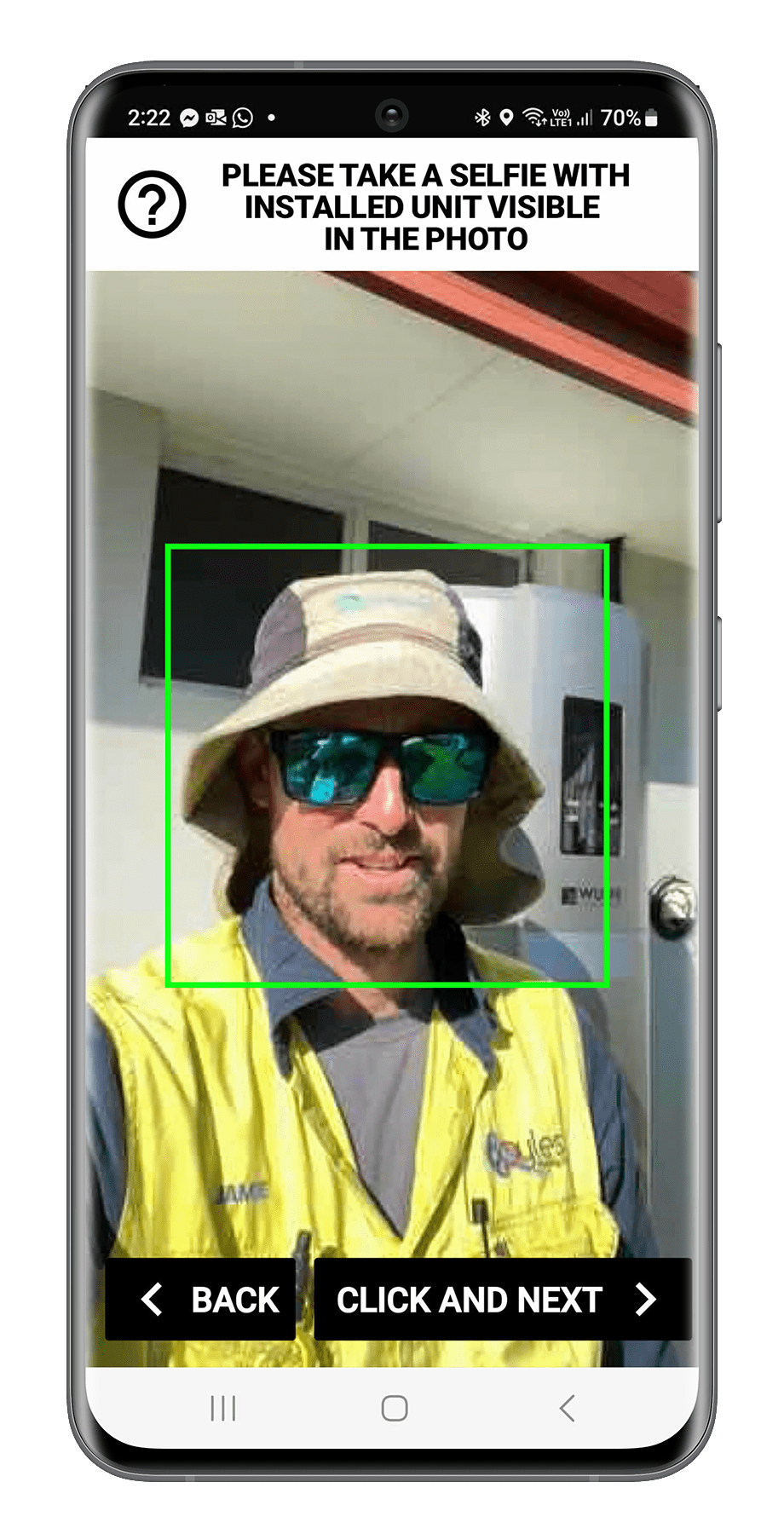 A man taking a selfie in front of an installed unit, wearing a wide-brimmed hat, sunglasses, a yellow vest, and a gray shirt, with instructions at the top to include the unit in the photo. The phone screen shows options to go back or proceed to the next step.