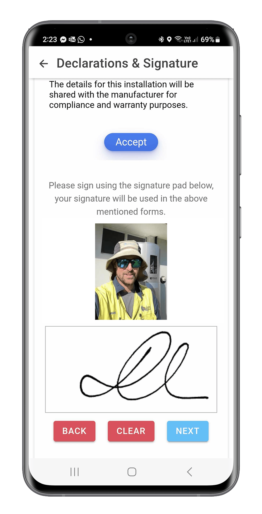 A smartphone screen showing a signature verification page with a profile photo of a man wearing sunglasses, a hat, and a yellow safety vest, above a signature box with a scribbled signature, and red and blue buttons labeled 'Back,' 'Clear,' and 'Next'.