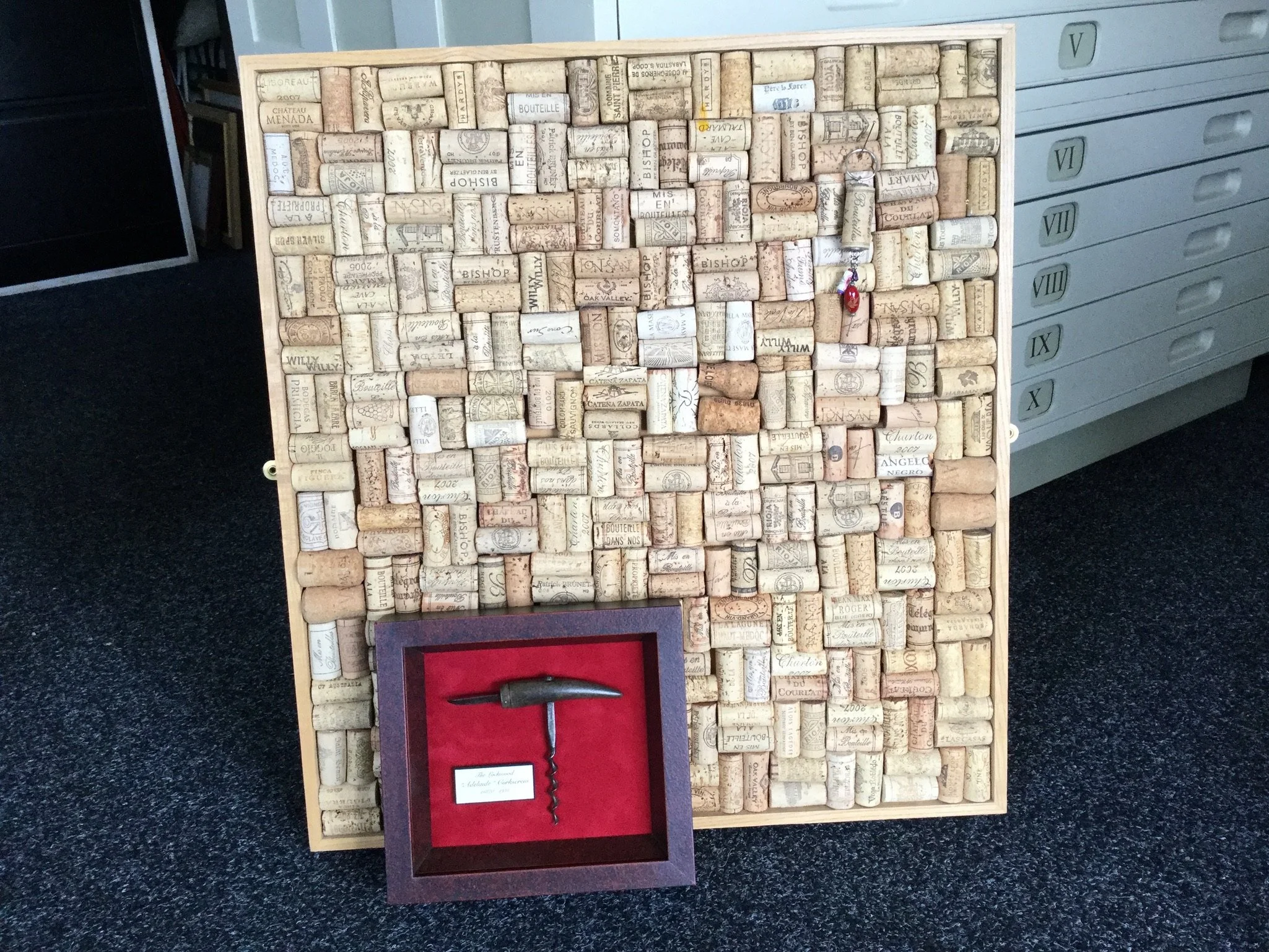 A cork board filled with wine corks and a keychain with a red ornament hanging from a cork. A framed corkscrew with a red interior is placed in front of the cork board.