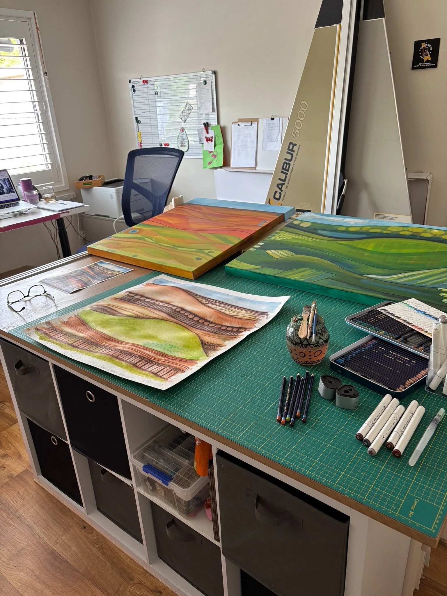 Art studio workspace with a green cutting mat on a table, displaying colorful paintings, markers, pens, and art supplies. In the background, a whiteboard, a chair, and an easel are visible.