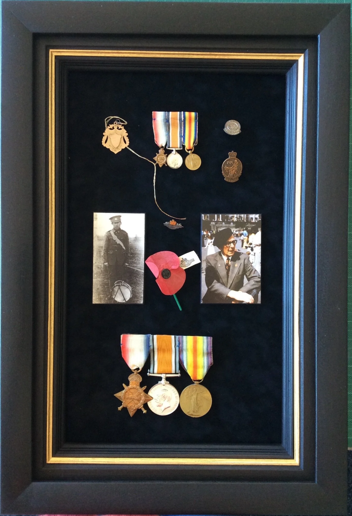 A shadow box display containing military medals, badges, a poppy flower, photographs, a pin, and pins, arranged on a dark background within a black frame with gold accents.