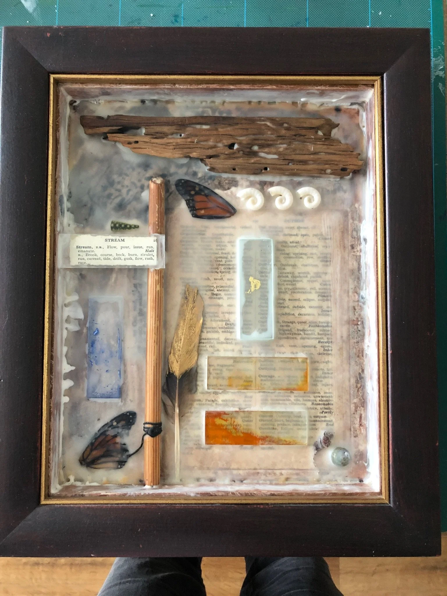 Mixed media art display inside a glass shadow box. Items include butterfly wings, dried wood, a feather, a matchstick, shells, and fragments of vintage paper and text.