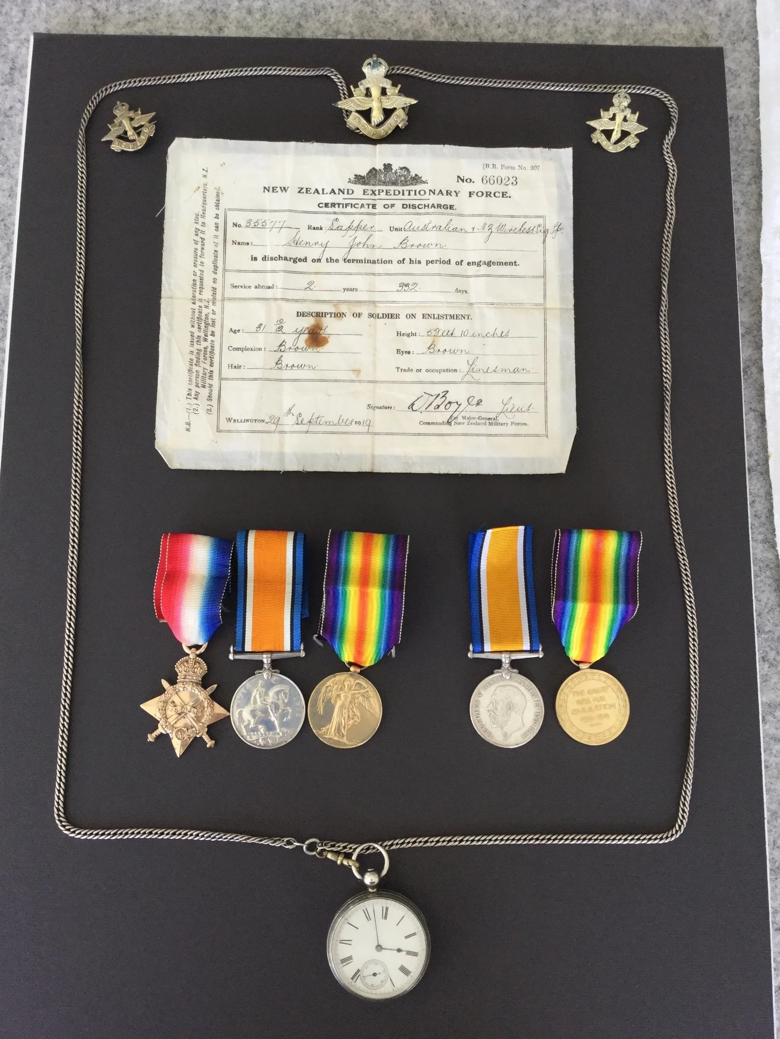 A display case containing a New Zealand Expeditionary Force discharge certificate, five military medals with colorful ribbons, and a pocket watch on a chain.