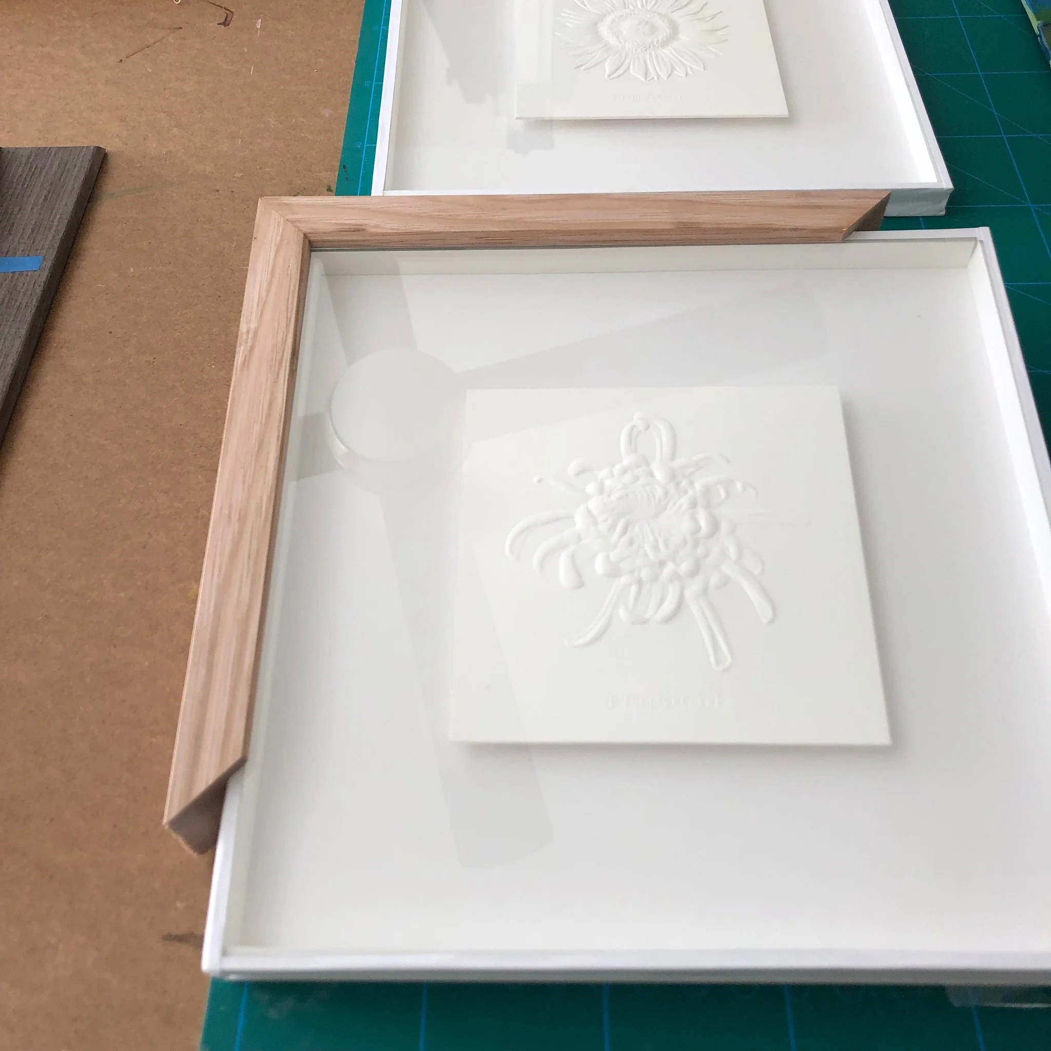 A white framed shadow box containing a white textured piece with a floral or abstract design, placed on a work surface.