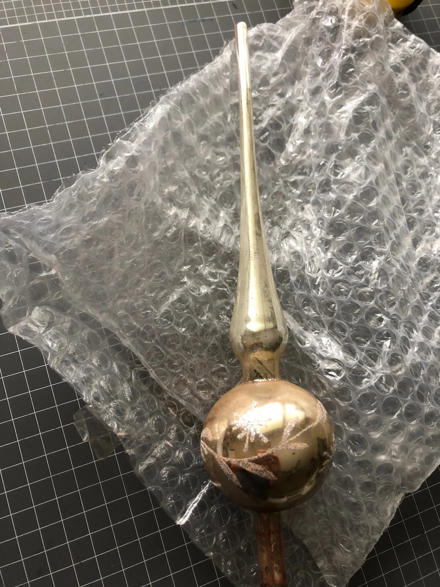 A gold-colored decorative finial with a spherical base and a tapered, elongated top, resting on bubble wrap on a grid-patterned surface.