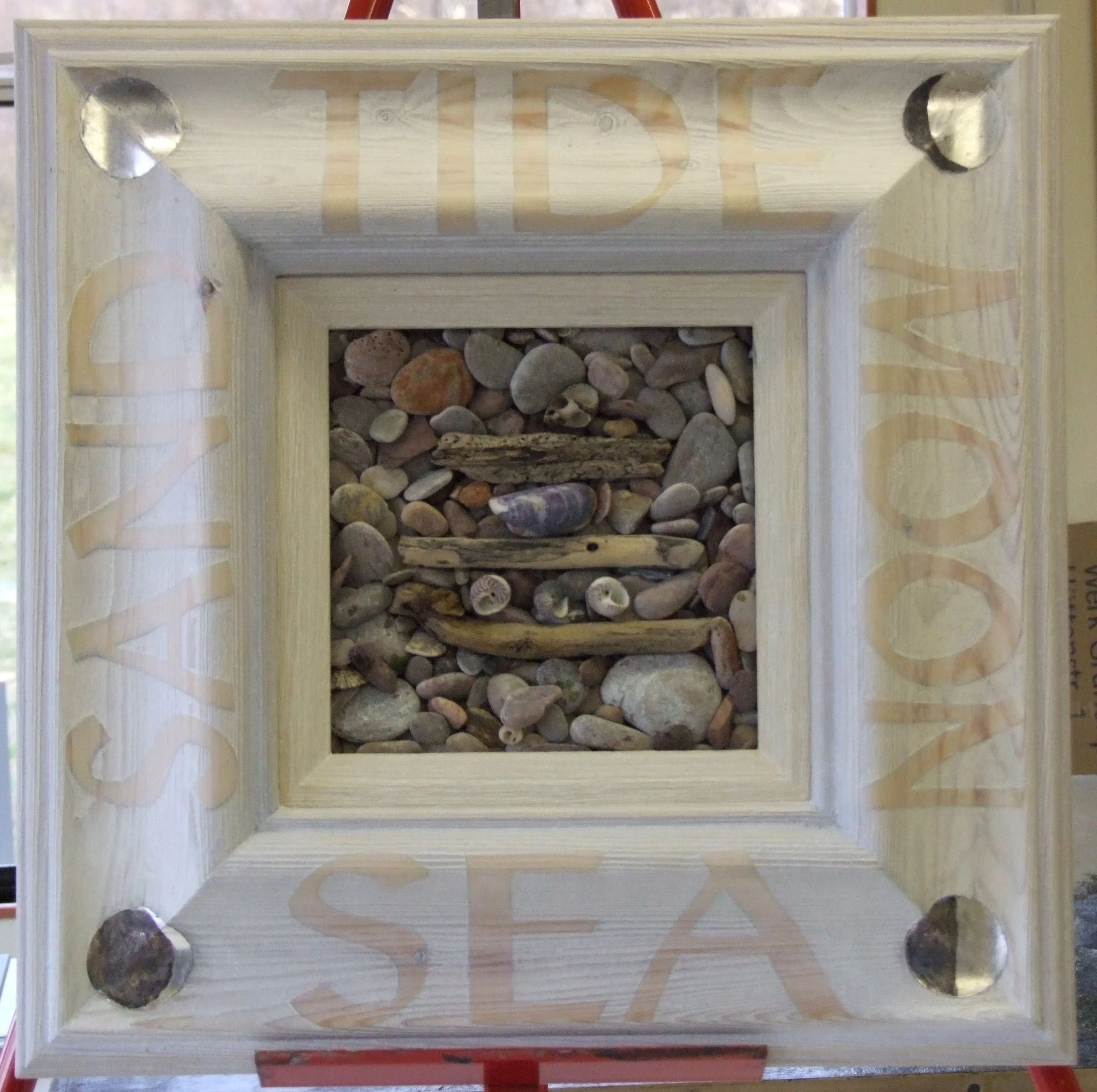 A square wooden shadow box with the word "SEA" carved into each side. Inside, the frame contains small stones, shells, and pieces of driftwood, creating a miniature coastal scene.