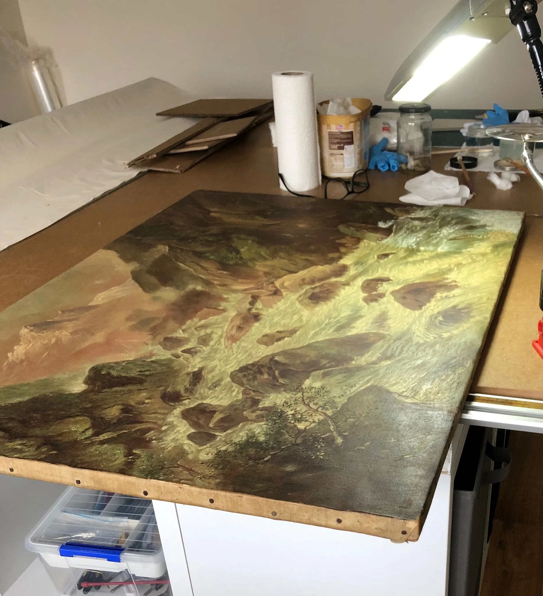 A landscape painting of a river cascading over rocks in a lush forest, placed on a worktable with art supplies and tools around.